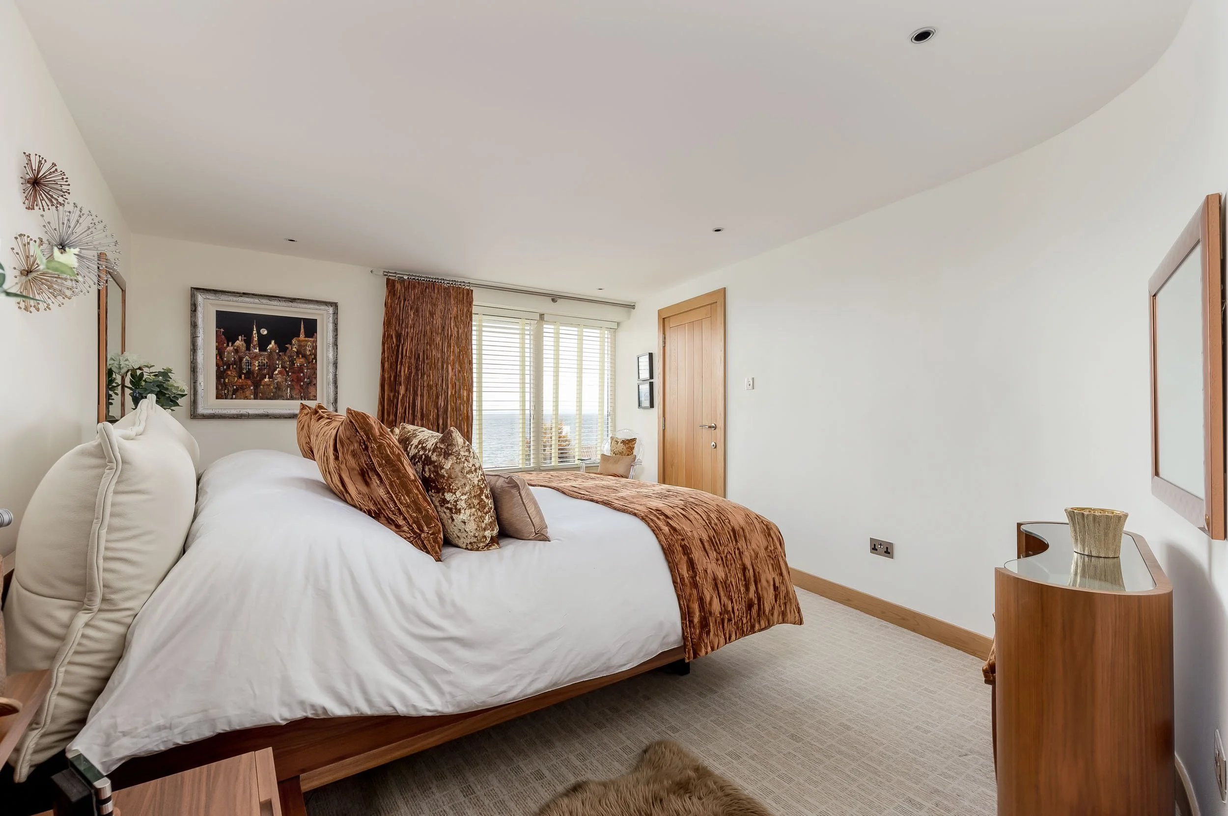 A bedroom with a large bed dressed in white sheets and brown velvet pillows, a window with orange curtains, a small chair by the window, wall art depicting a cityscape, and a wooden cabinet with a decorative vase on top.