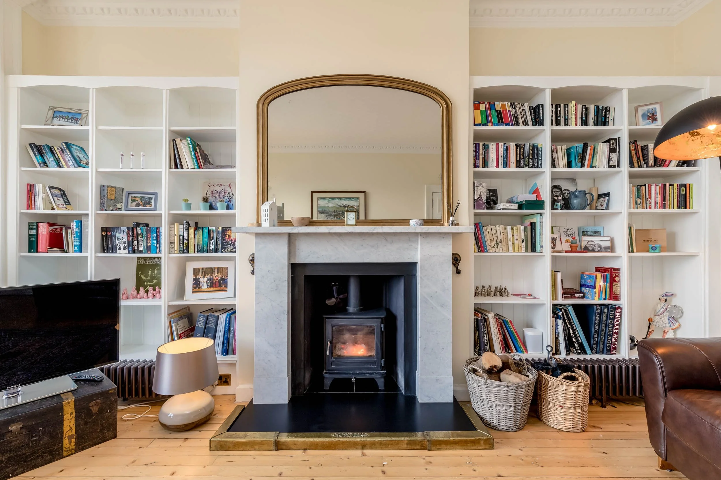 Living room with white built-in bookshelves flanking a fireplace, which has a gold-framed mirror above it, and a variety of books and decorative items on the shelves and mantel. There is a television and a lamp on the left, and two baskets of logs on