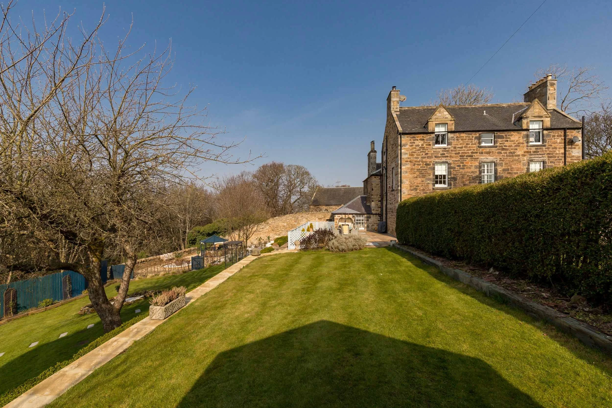 A well-maintained garden with a lush green lawn, a pathway, a leafless tree, and a large stone house with multiple windows and chimneys under a clear blue sky.