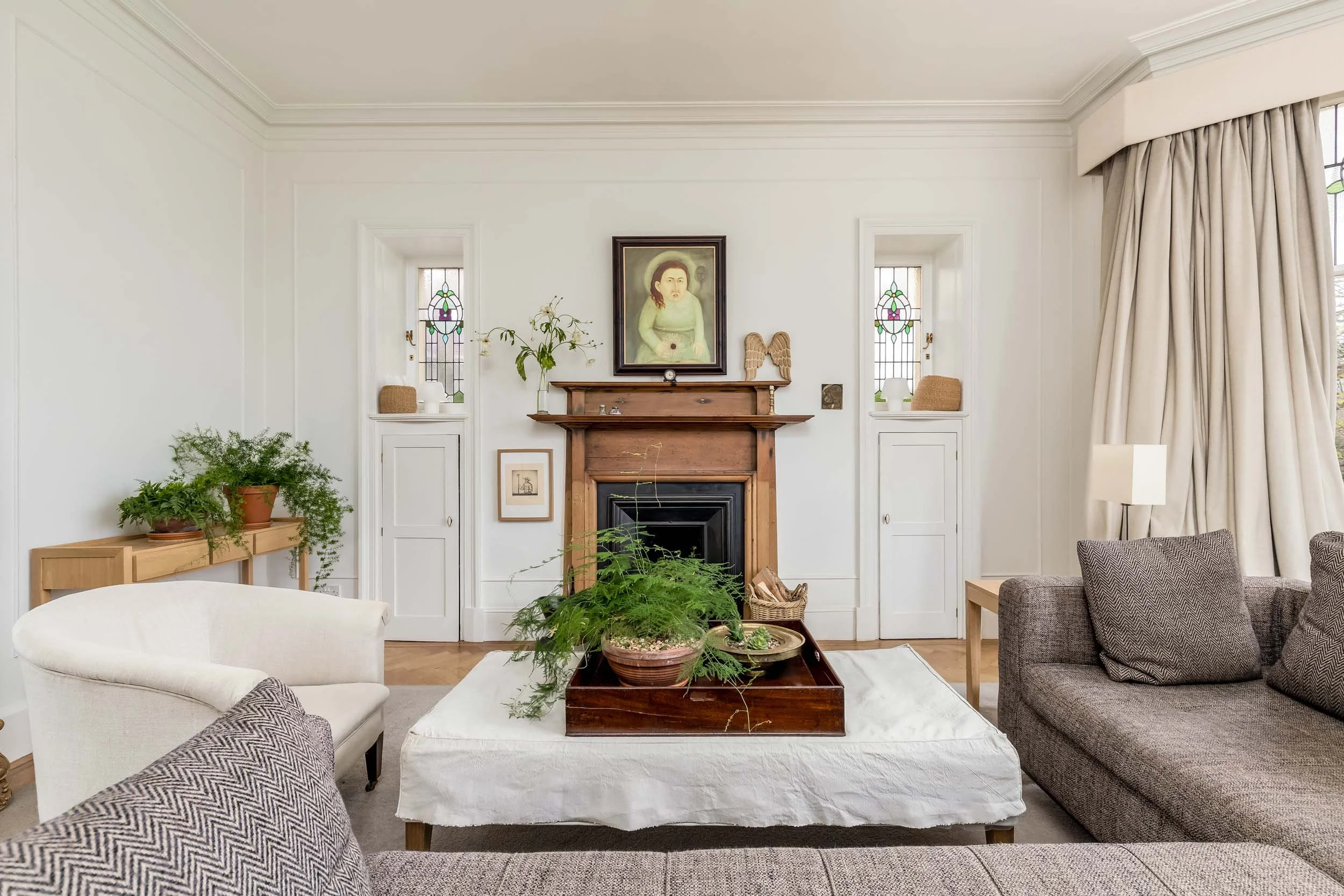 Living room with a fireplace, stained glass windows, and neutral furniture, featuring plants and artwork.