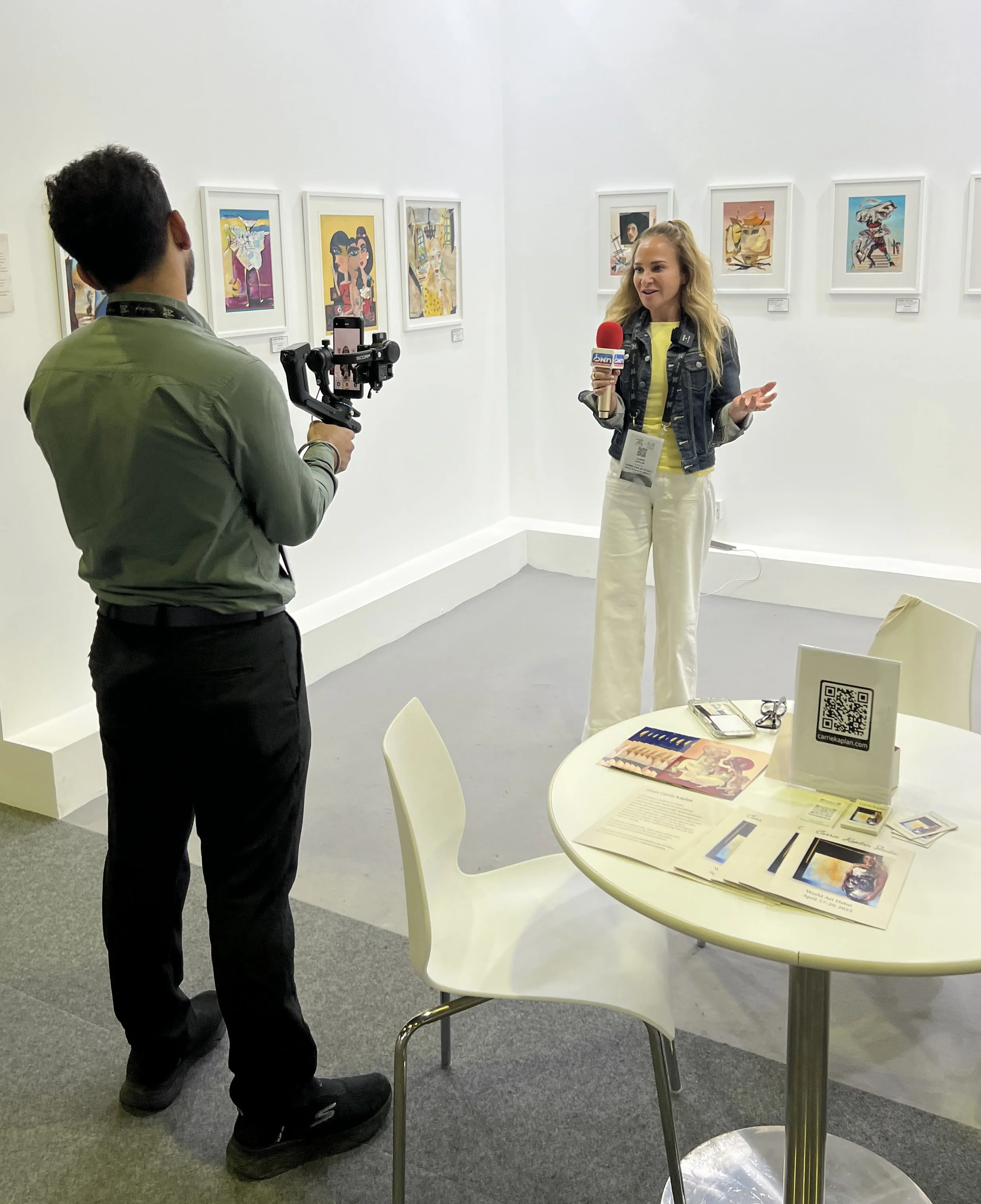 A woman is being interviewed with a microphone in an art gallery, while a man records her with a camera stabilizer. The gallery wall behind her displays colorful artworks.