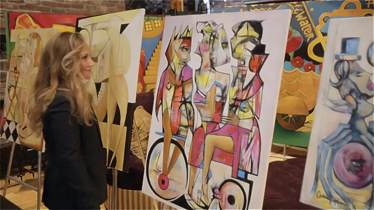 A woman with long blonde hair, dressed in a black blazer, looks at colorful abstract paintings in an art gallery.