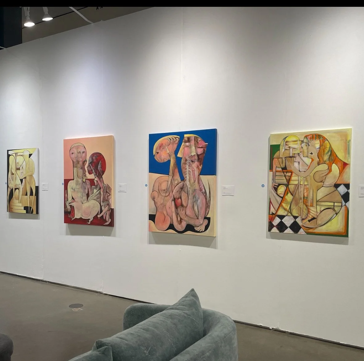 An art gallery displays four abstract paintings featuring distorted human figures and faces in various color palettes, hung on a white wall.