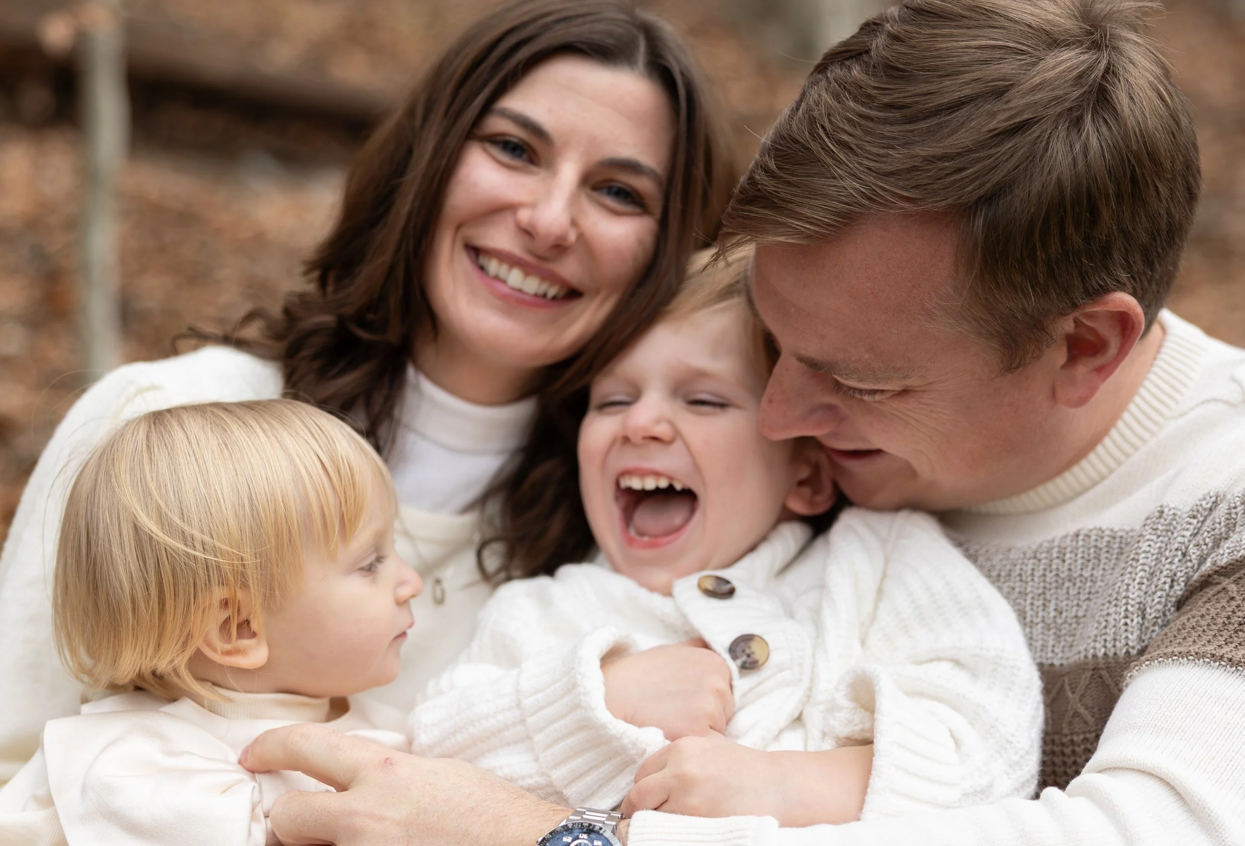 A happy family of four, including a mother, father, and two young children, enjoying a moment together outdoors. The children are laughing and the parents are smiling warmly.