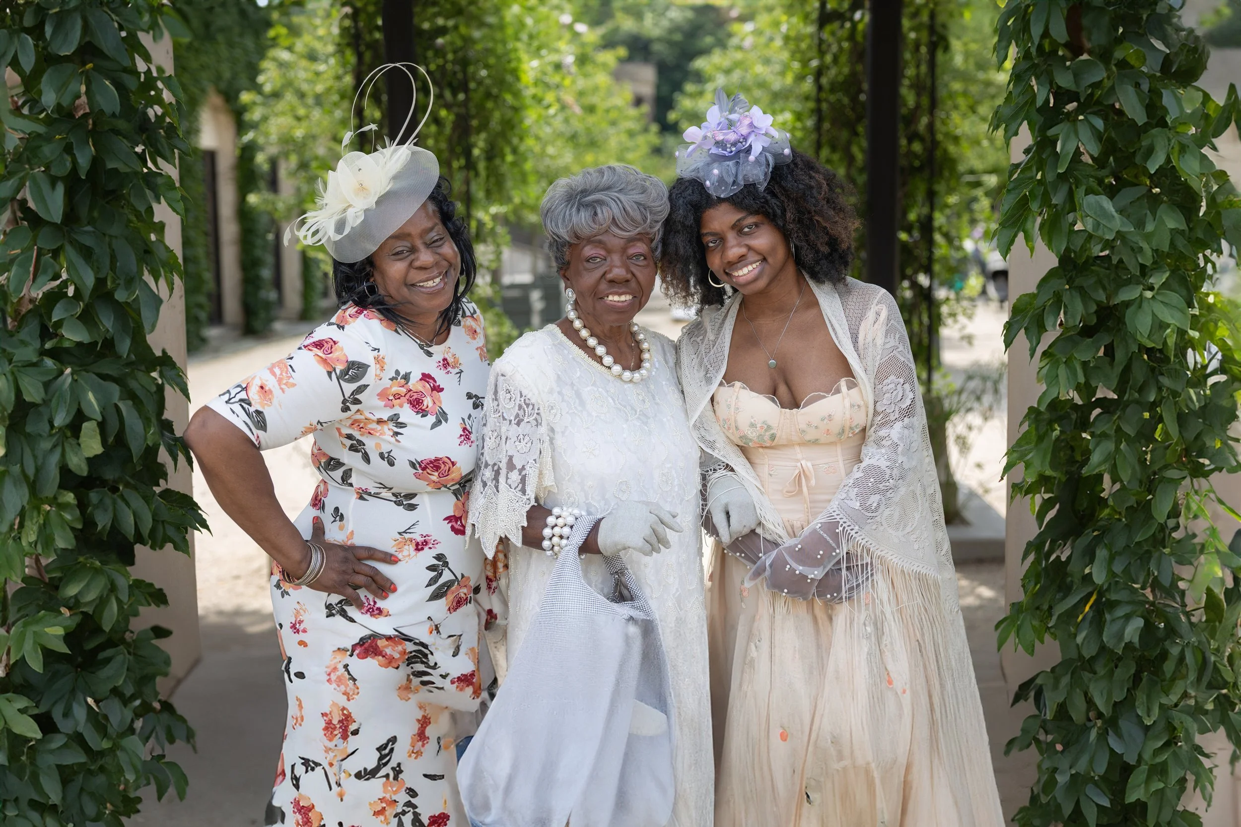 Three women dressed in vintage-style clothing, smiling and posing outdoors under a green arbor.