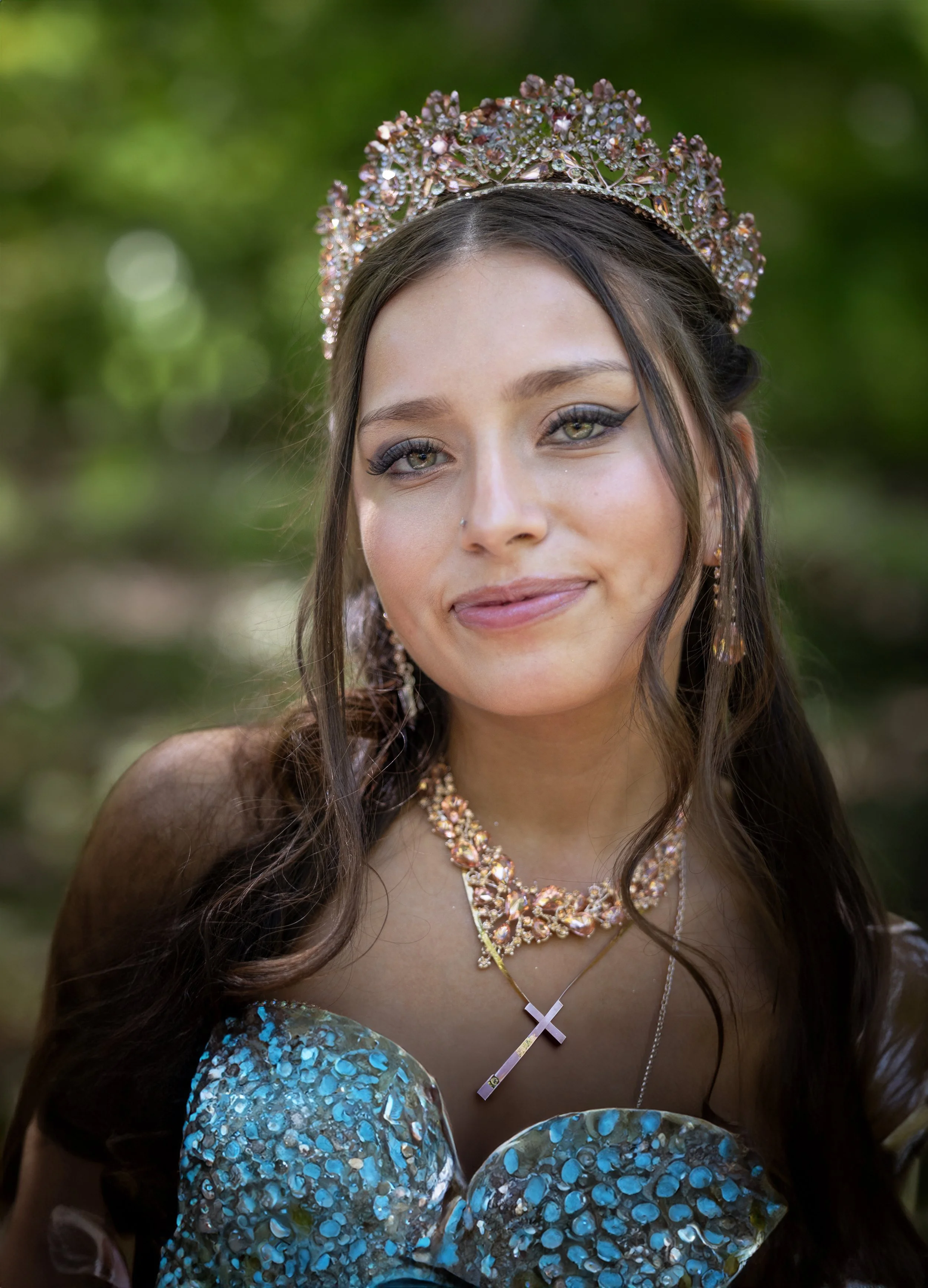 Young woman with long brown hair, wearing a jeweled crown, and dressed in an elaborate gown with a shimmer pattern. She has makeup with winged eyeliner, and wears a necklace with a cross pendant and sparkling earrings. The background is blurred green