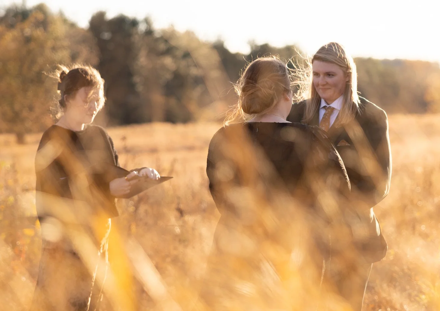 Three women standing in a field during sunset. One woman is on the left holding a notepad or phone, while two women on the right are conversing. All are dressed in formal or business attire. The background includes trees and a warm, golden light.