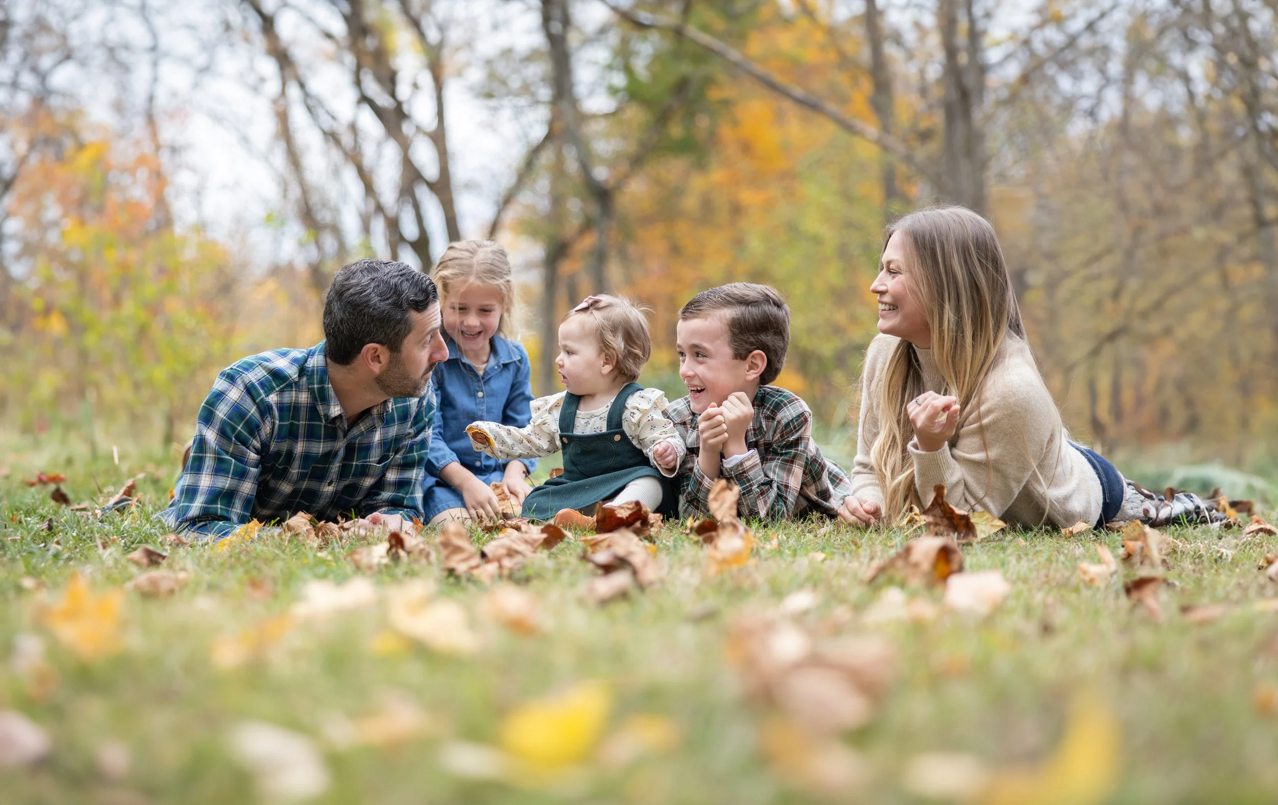 A family of six lying on the grass in a park during fall, laughing and playing together, surrounded by colorful autumn leaves and trees.