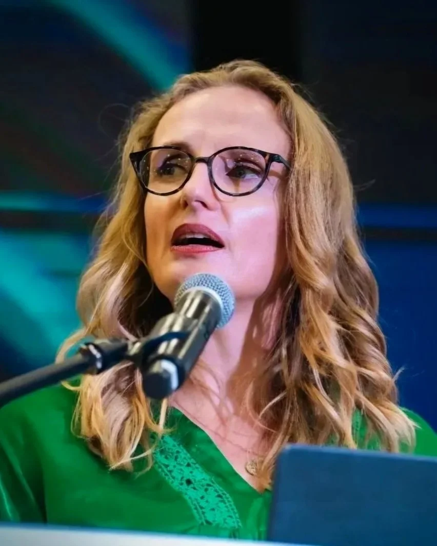 A woman with long wavy hair and glasses speaking into a microphone, wearing a green top, against a colorful background.