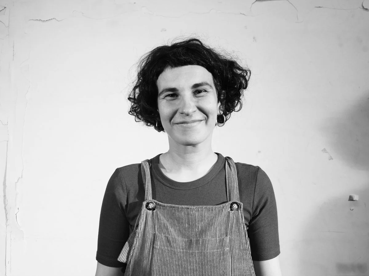 Schwarz-weiss-Fotografie einer lächelnden, jüngeren Frau mit kurzem, lockigem Haar, die eine Cord-Overall trägt, vor einem weissen Hintergrund.