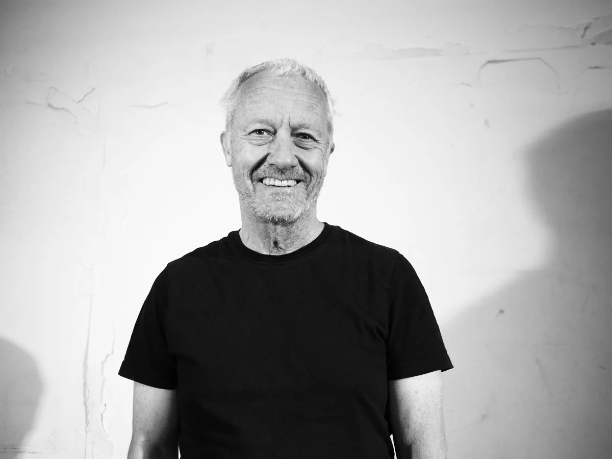 Ein lächelnder Mann mit grauem Haar und leichtem Bart, trägt ein schwarzes T-Shirt, steht vor einer hellen Wand mit leichten Rissen, schwarz-weiss Foto.