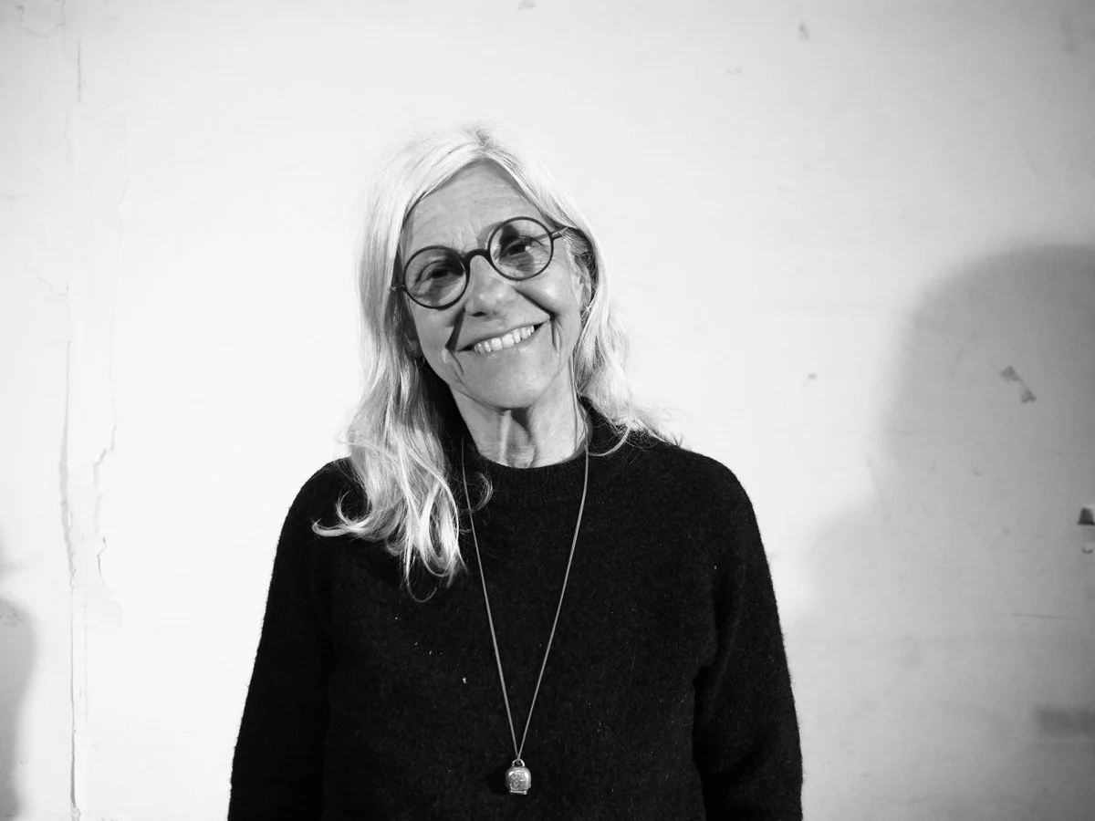 Ein Frau mit langen weissen Haaren, runder Brille und schwarzem Pullover, lächelnd, steht vor einem weissen Hintergrund.