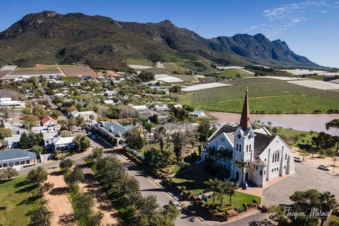 A picturesque small town with a prominent white church featuring a tall steeple, surrounded by green trees, with mountains in the background and vineyards in the distance on a clear day.