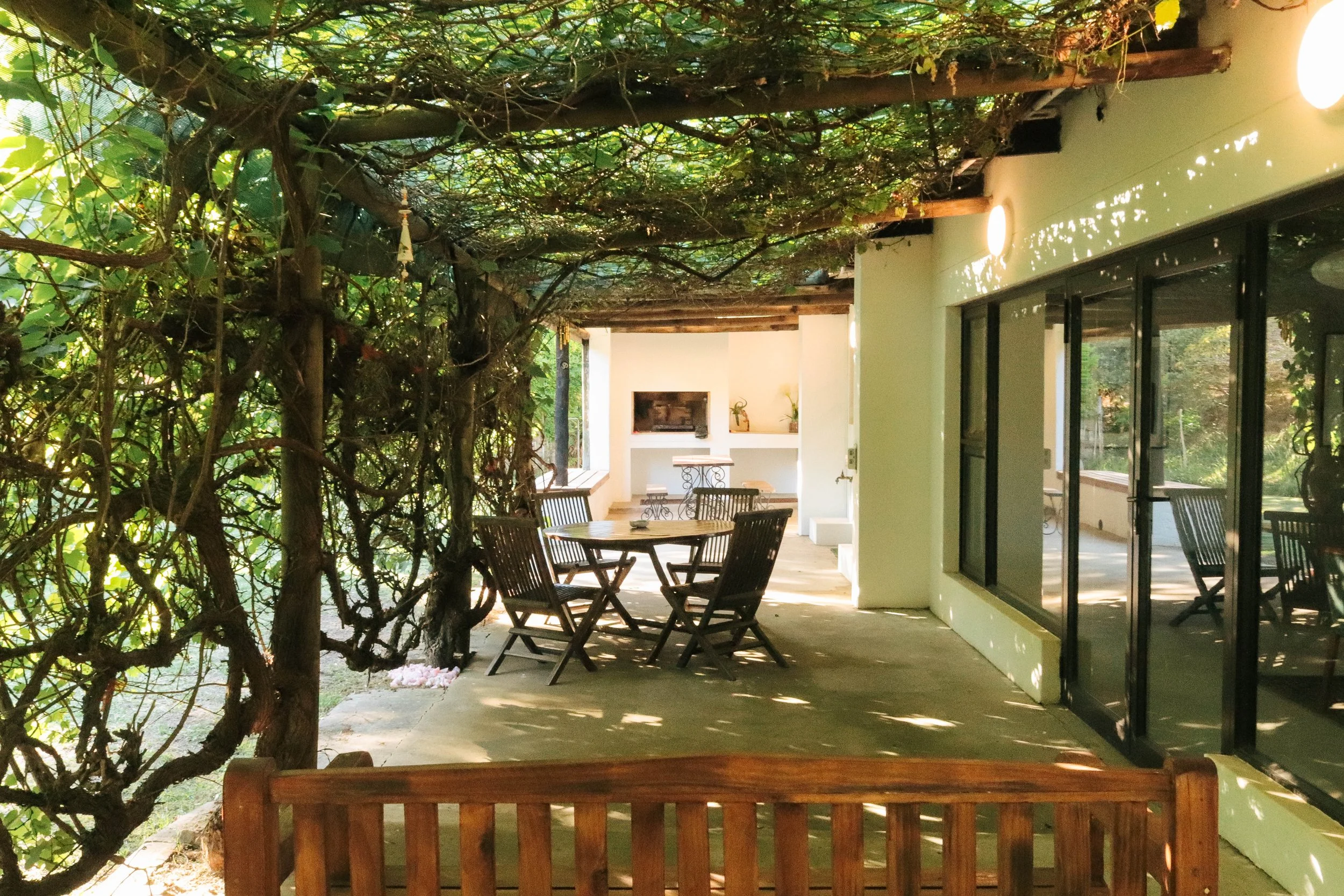 Covered outdoor veranda with wooden chairs and a table, adjacent to a building with glass sliding doors, shaded by a vine-covered roof.