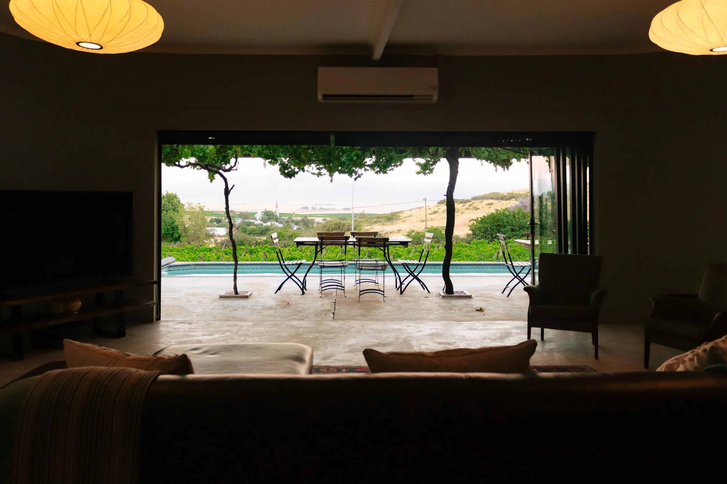 Interior living room with large open sliding glass door leading to an outdoor patio with a view of trees, hills, and a pool. Outdoor table and chairs are visible.
