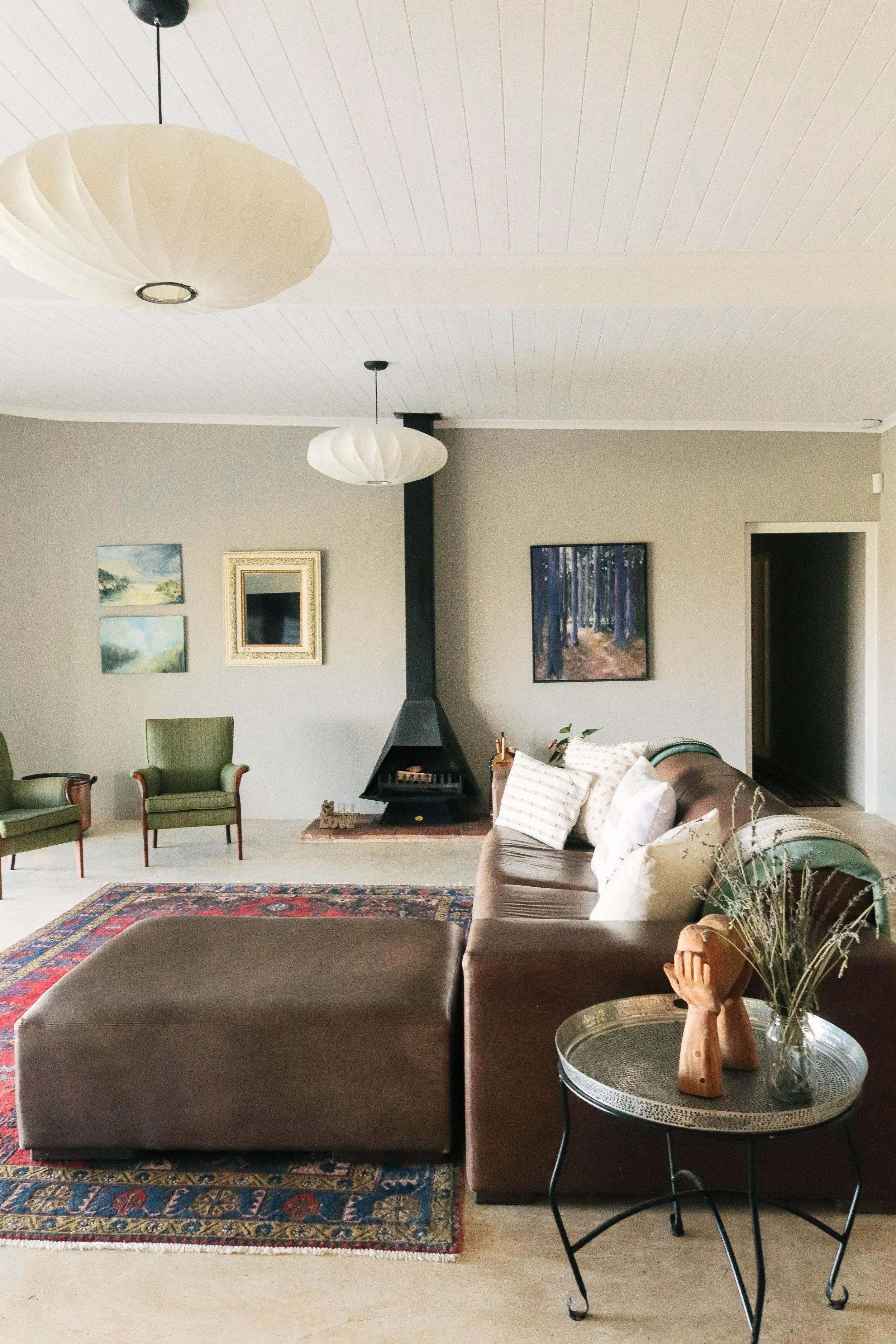 Living room with a brown sectional sofa, green armchairs, a fireplace, framed artwork on the walls, a patterned area rug, and a small round side table with decorations.