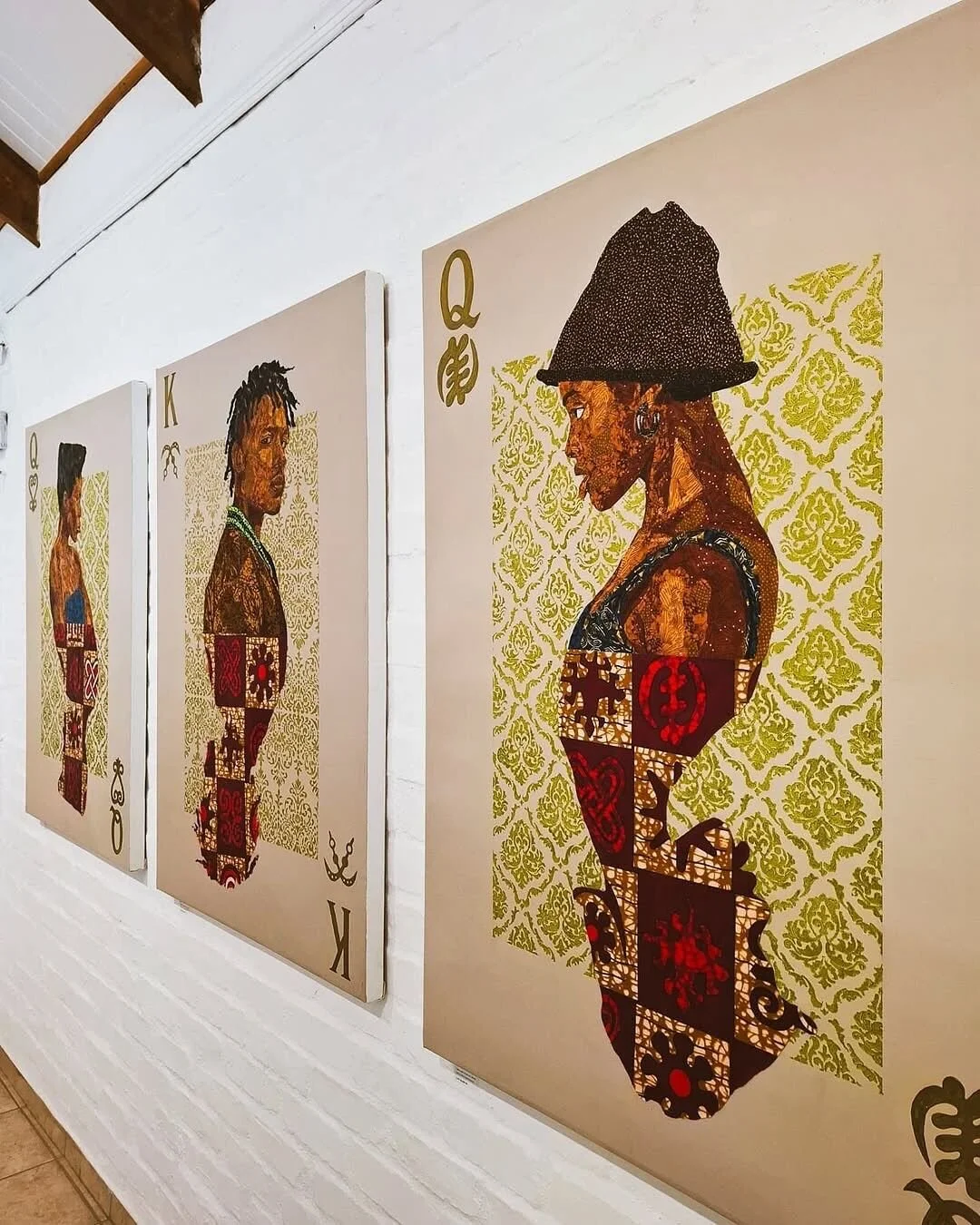 Three playing card paintings on a white brick wall, depicting women in profile with tribal-inspired patterns and textures, each representing different card ranks: Queen, King, and Queen, with detailed backgrounds and accessories.