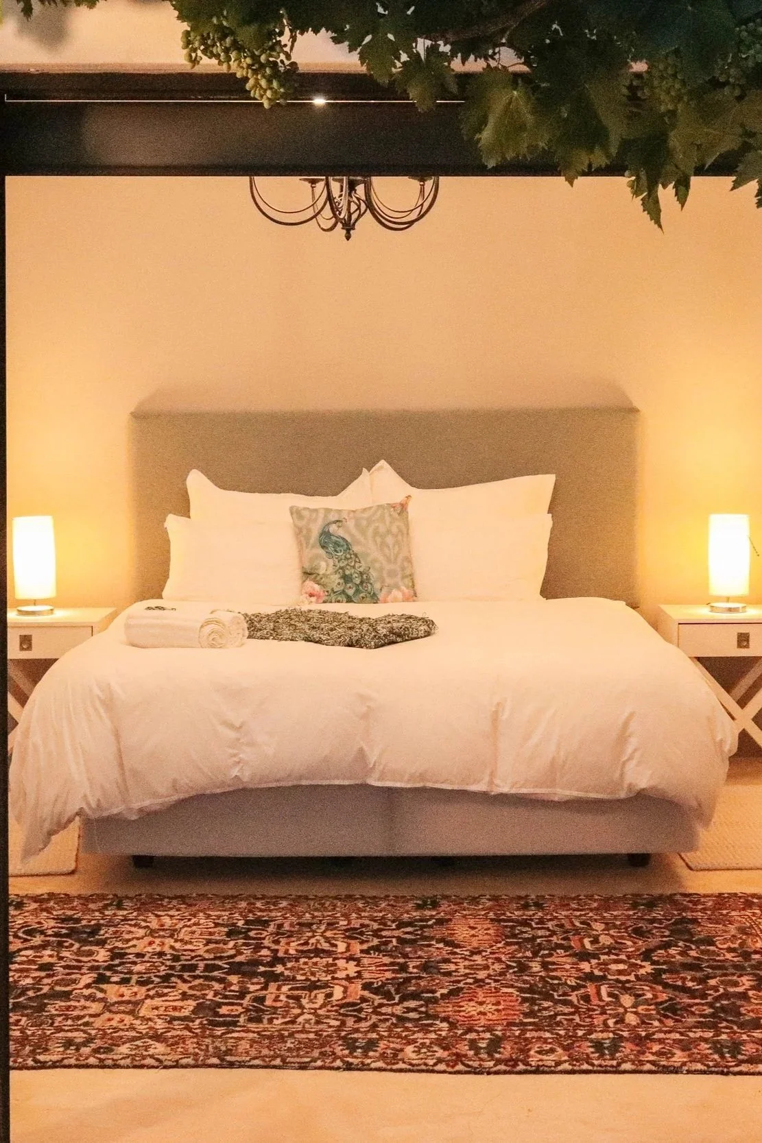 A neatly made bed with white bedding, a peacock-themed decorative pillow, two rolled towels, a patterned cloth, and two bedside lamps emitting warm light, set against a beige wall with a headboard and a rug in front.