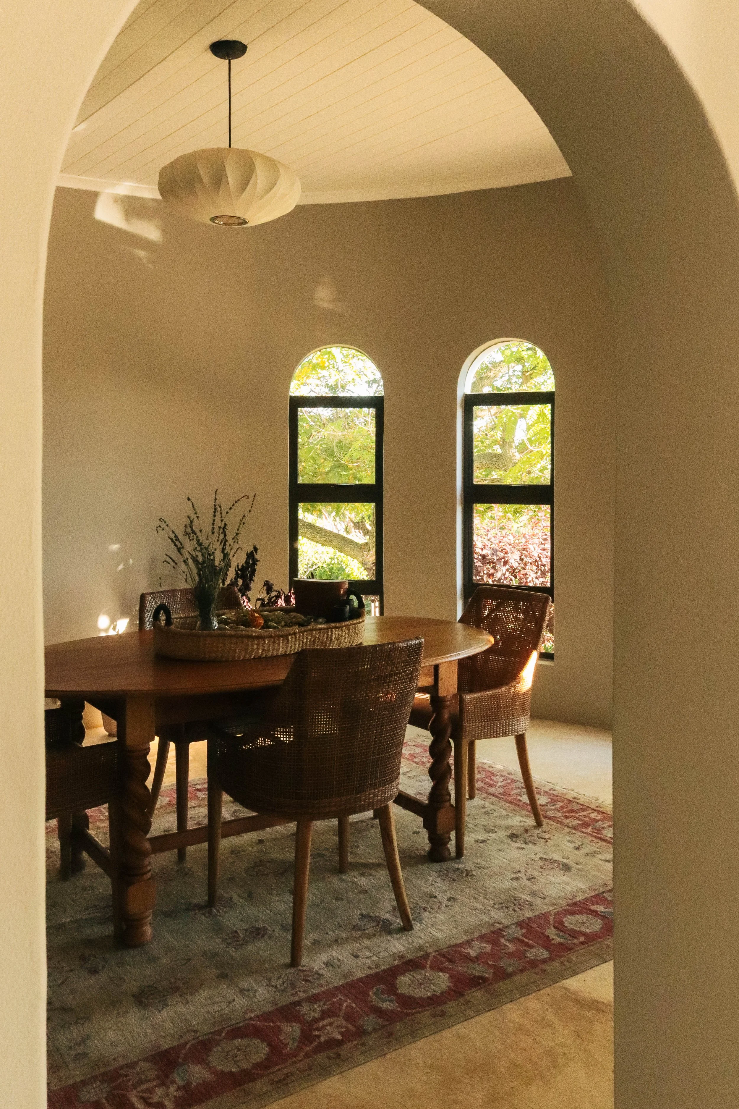 A cozy dining area with a wooden table and four woven chairs, located near two large arched windows showing trees outside. There is a hanging ceiling light and a decorative rug on the floor.