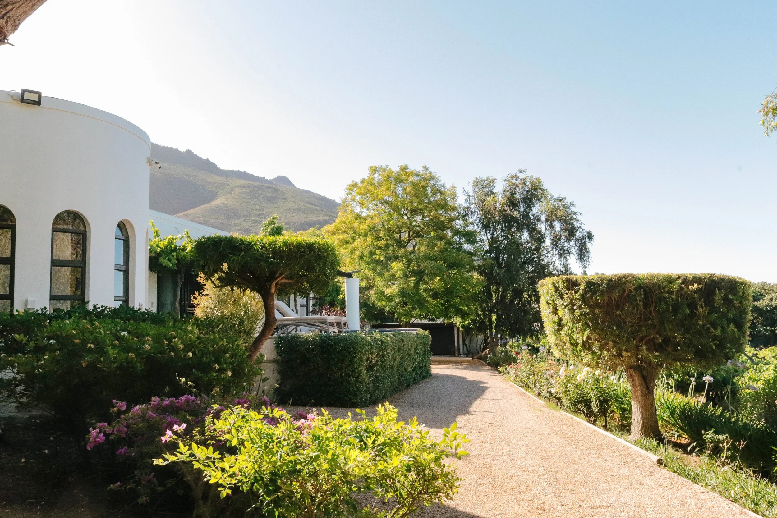 A bright outdoor garden scene with a white modern building partially visible on the left, a gravel pathway, and various trimmed trees and bushes, with hills in the background under a clear sky.