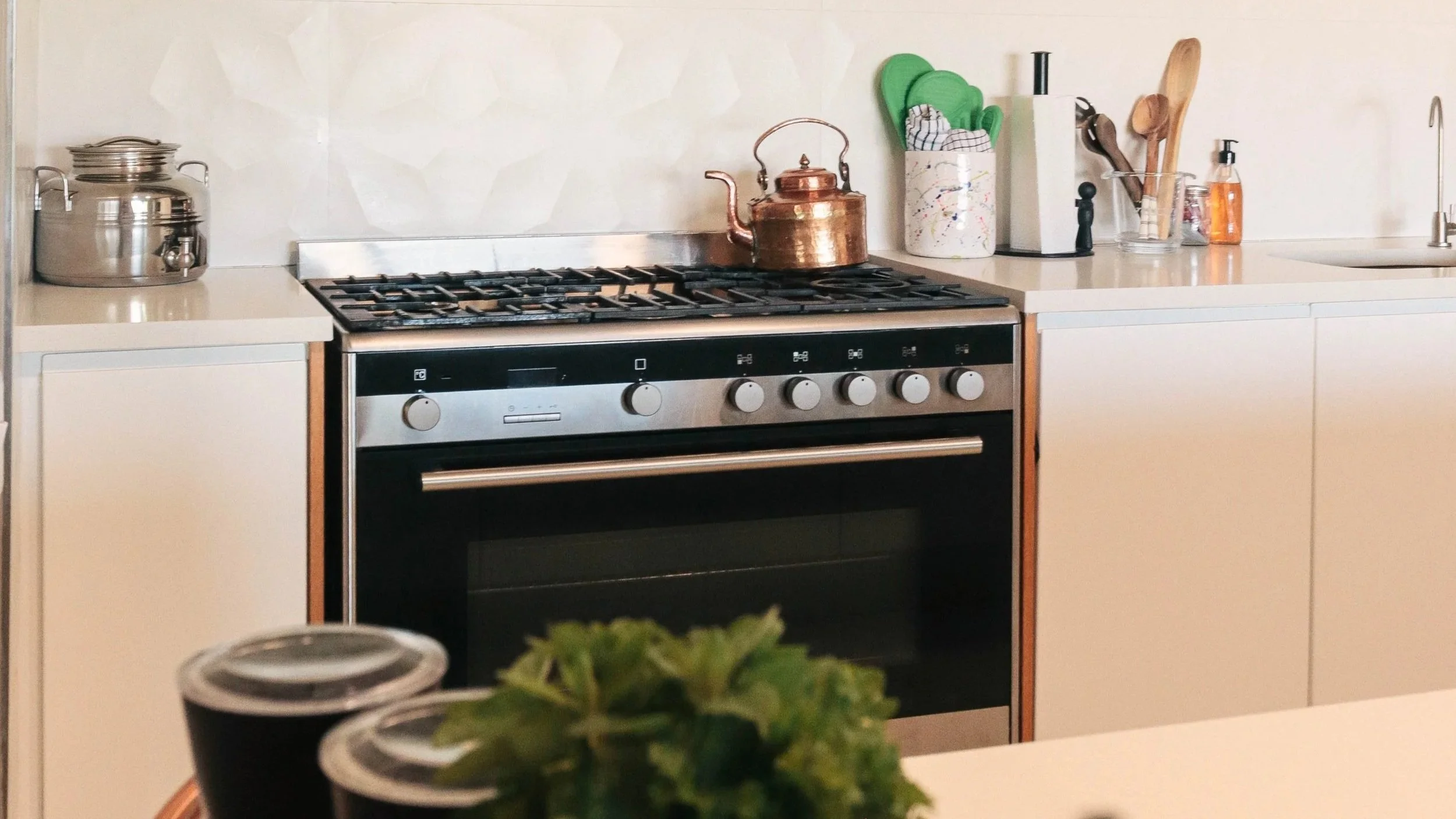 Kitchen with a stove, copper kettle, and countertop items like herbs, cups, and utensils.