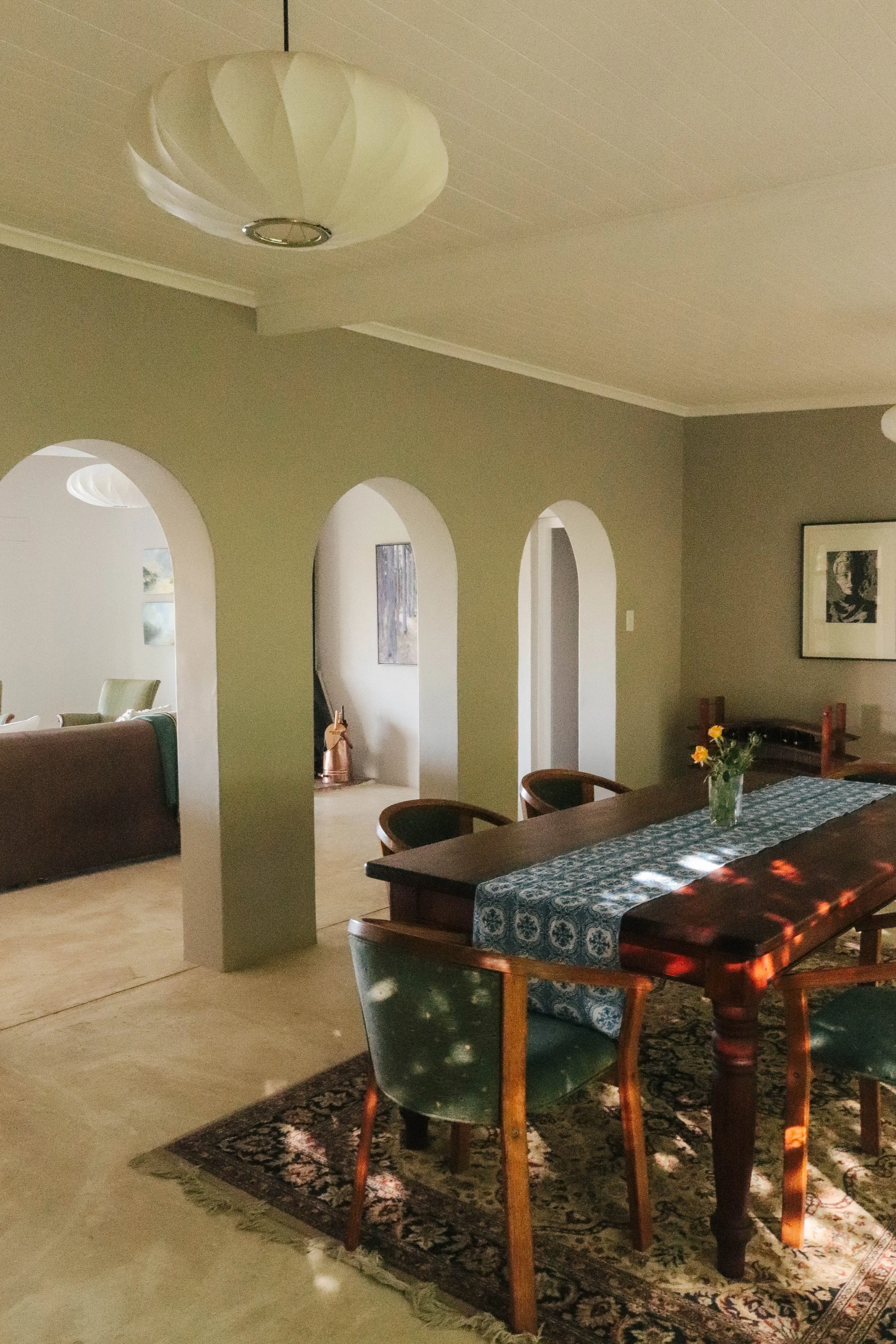Dining room with a large wooden table, green chairs, a floral table runner, and a small vase with yellow flowers, illuminated by sunlight.