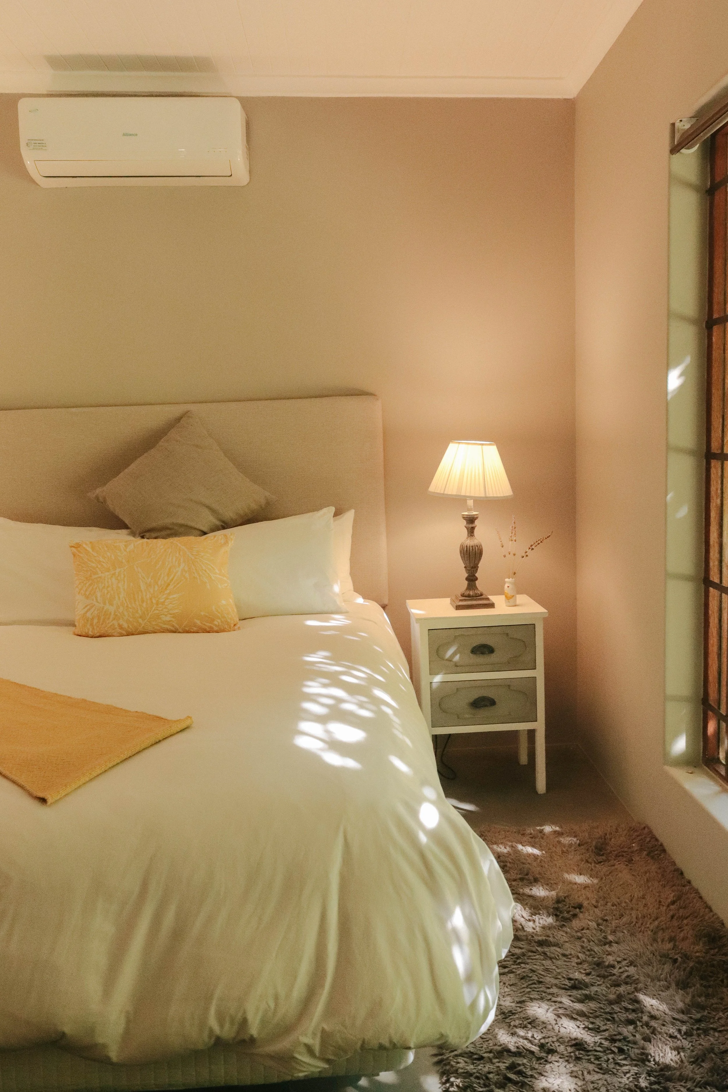 A cozy bedroom featuring a bed with a beige headboard, white bedding with yellow and gray pillows, a bedside table with a lamp, and a window with wooden framing allowing natural light to filter in.