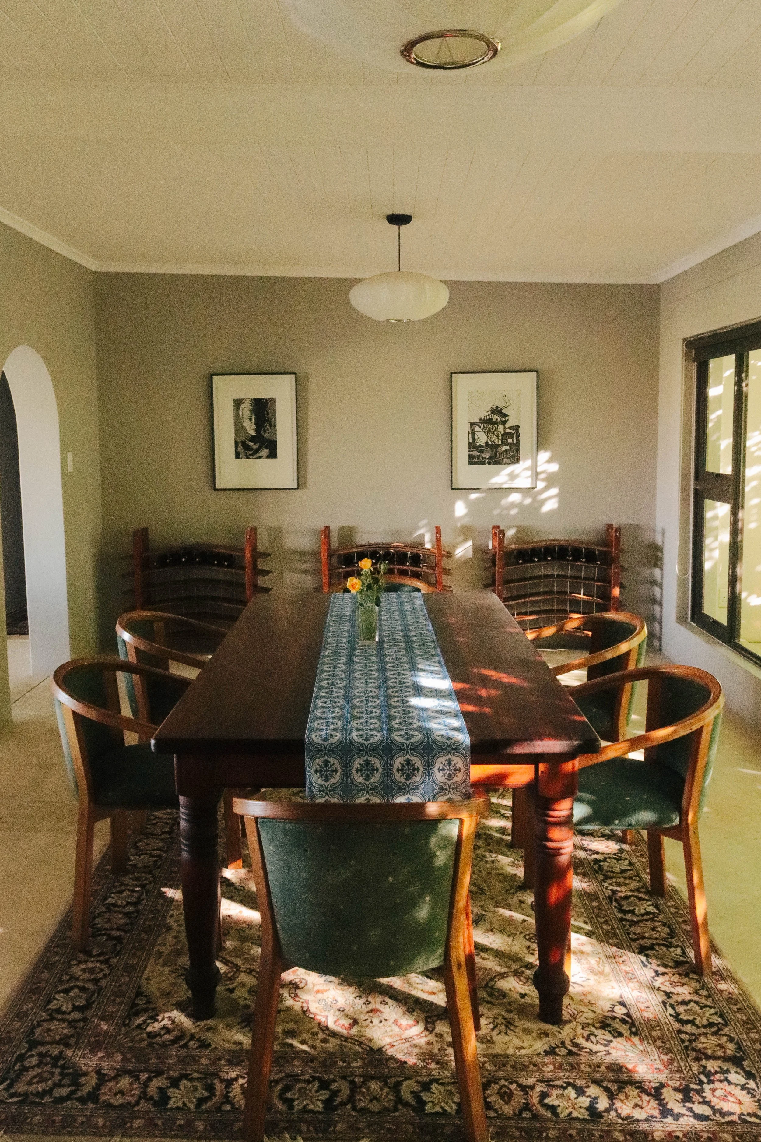 A dining room with a wooden table, six chairs with green upholstery, a patterned table runner, a small vase with flowers, framed artwork on the wall, and sunlight coming through a window.