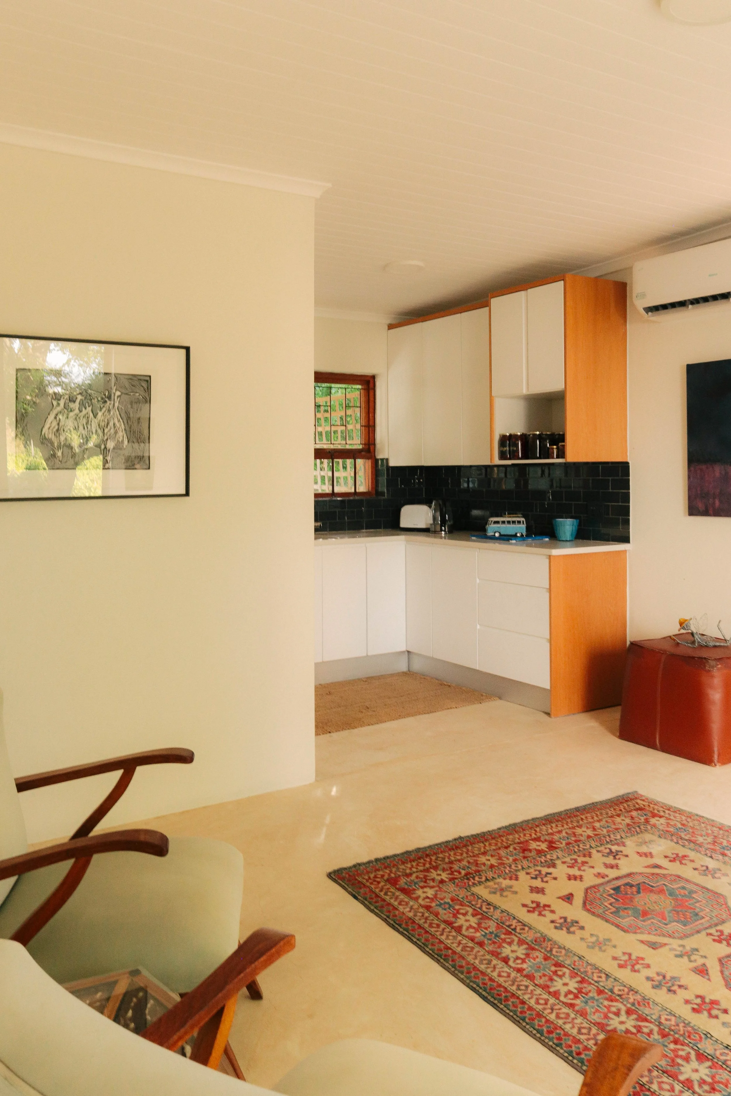 A cozy living space with a partial view of a kitchen, a decorative rug on the floor, a wooden chair with light-colored cushions, and framed artwork on the wall.