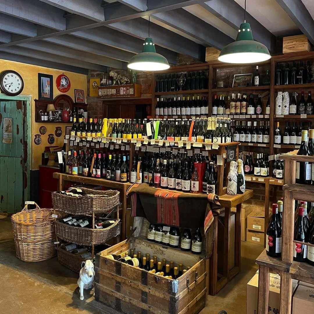 A cozy wine store displaying shelves filled with wine bottles, baskets of wine bottles on the floor, and vintage decor. Two green pendant lights hang from the ceiling, illuminating the space.