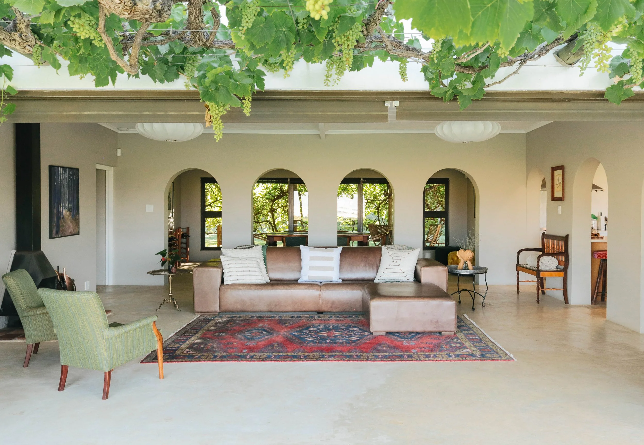 Living room with a beige leather sectional sofa, green striped armchairs, and a patterned area rug. Windows and arched doorways show a view of trees outside. The ceiling has white beams and light fixtures, and the space contains various decorative items and furniture pieces.