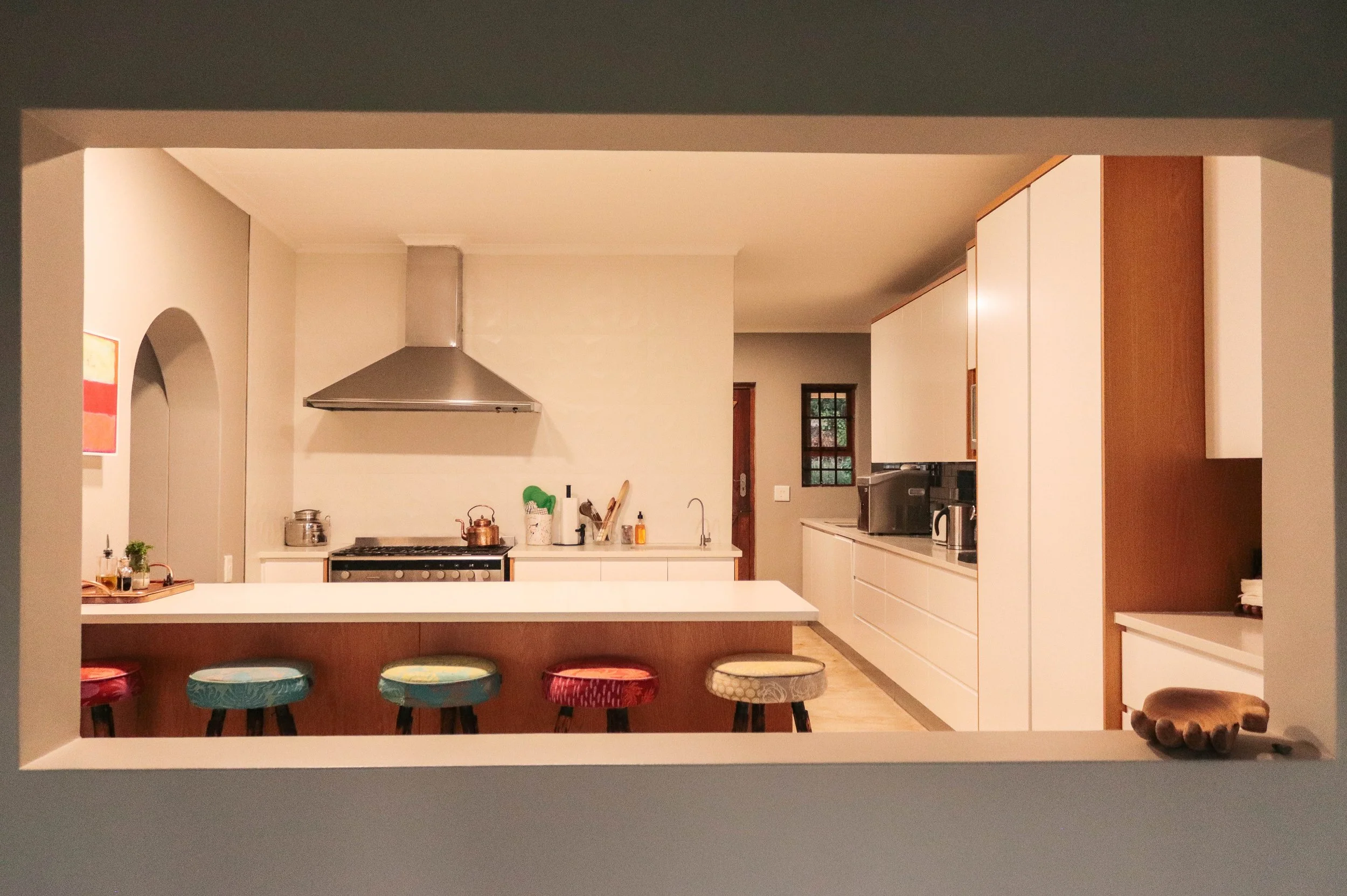 View through a window showing a modern kitchen with a white countertop, colorful barstools, appliances, and cabinetry.