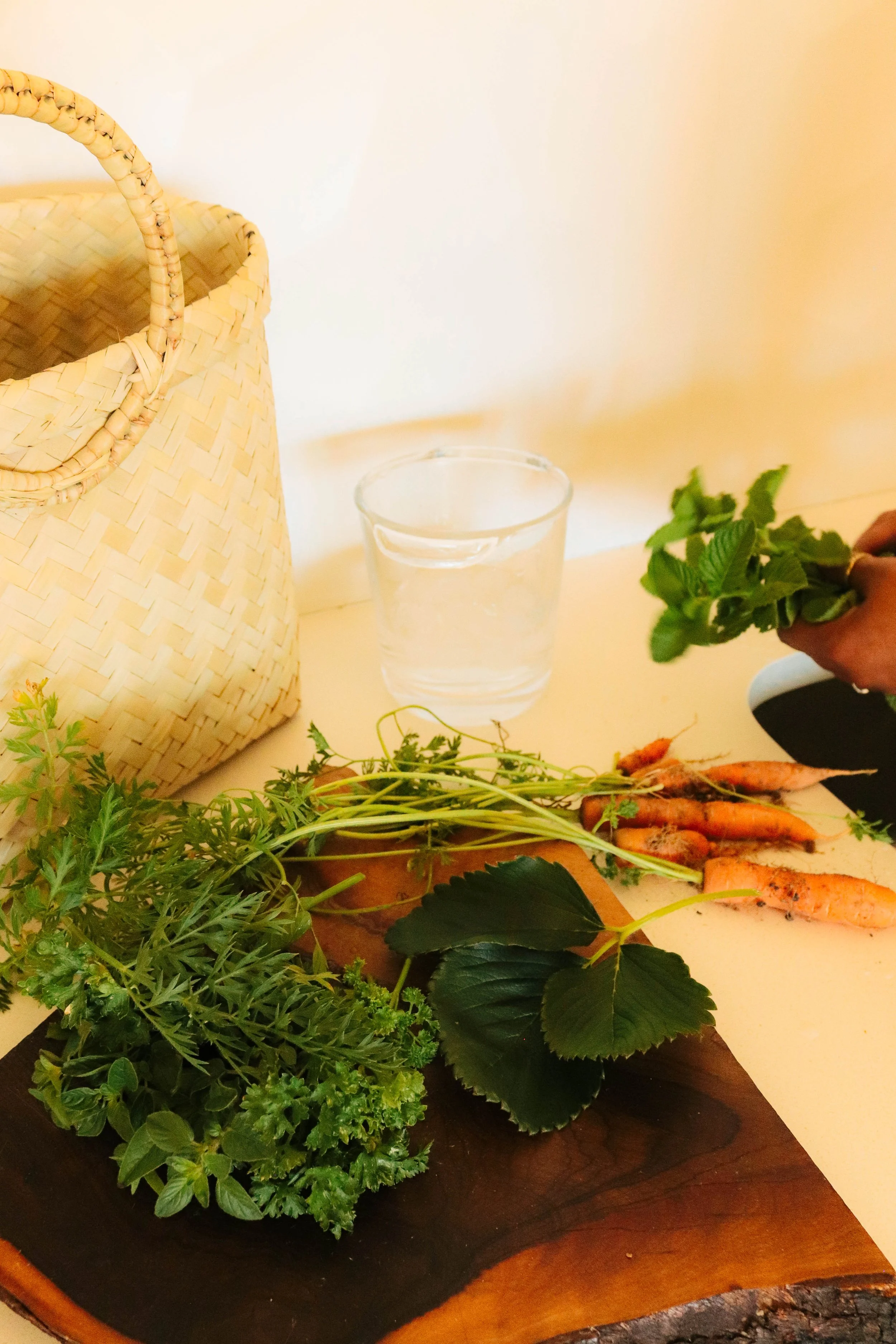 Fresh herbs and carrots on a wooden cutting board, with a glass of water and a woven basket nearby.