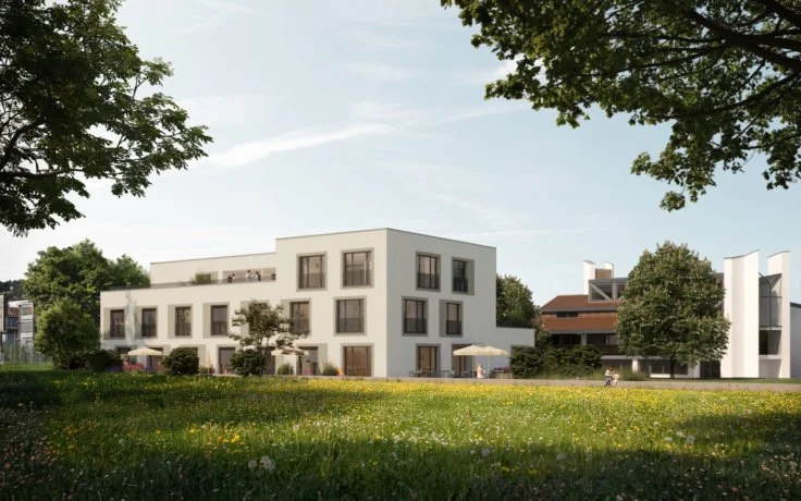 Moderner, weißer Wohnkomplex mit mehreren Etagen, umgeben von Grünflächen und Bäumen, bei sonnigem Wetter.