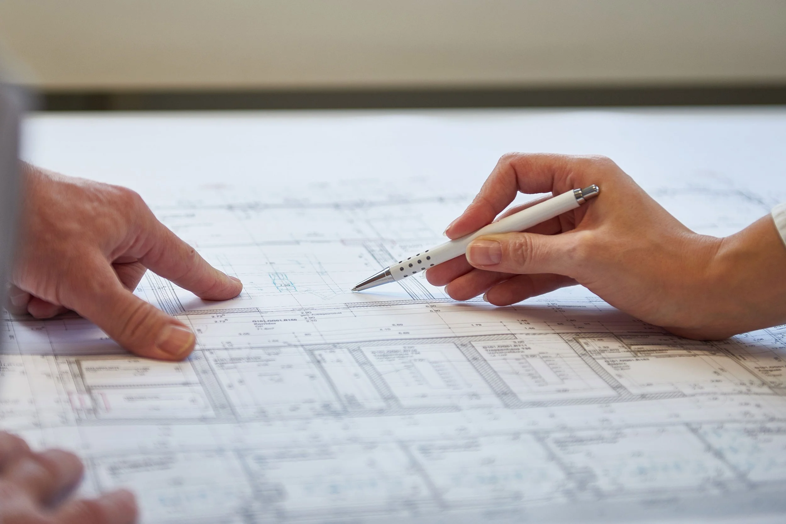 Nahaufnahme von zwei Personen, die eine technische Zeichnung auf einem Tisch besprechen, eine Person zeigt auf die Zeichnung und die andere hält einen weißen Kugelschreiber.