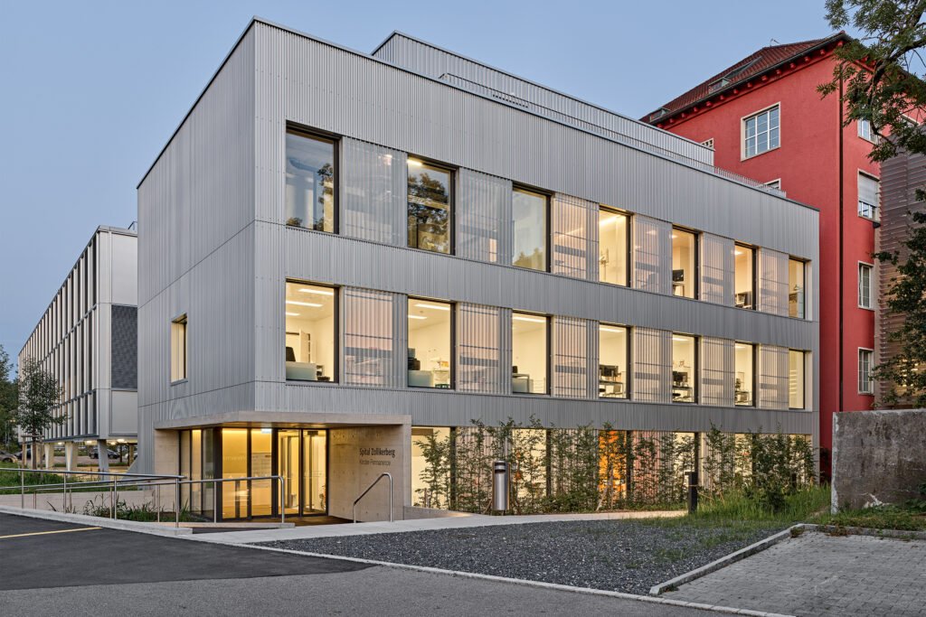 Moderne weiße Bürofassade mit großen Fenstern, umgeben von anderen Gebäuden, bei Dämmerung, mit beleuchteten Innenräumen.