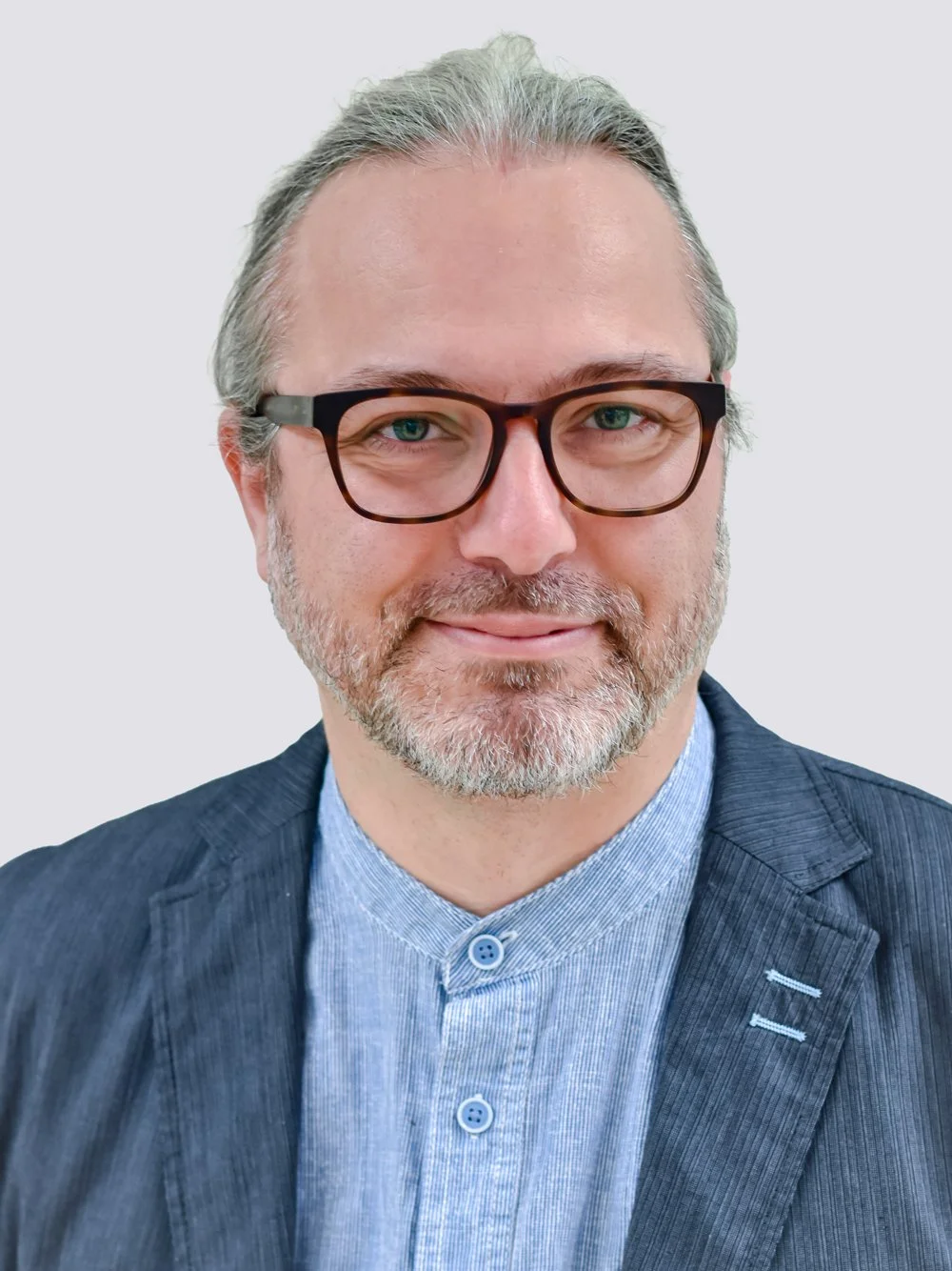 Porträt eines Mannes mit grauem Haar, Brille und Bart, trägt eine blaue Hemd und einen dunklen Blazer, vor hellem Hintergrund.