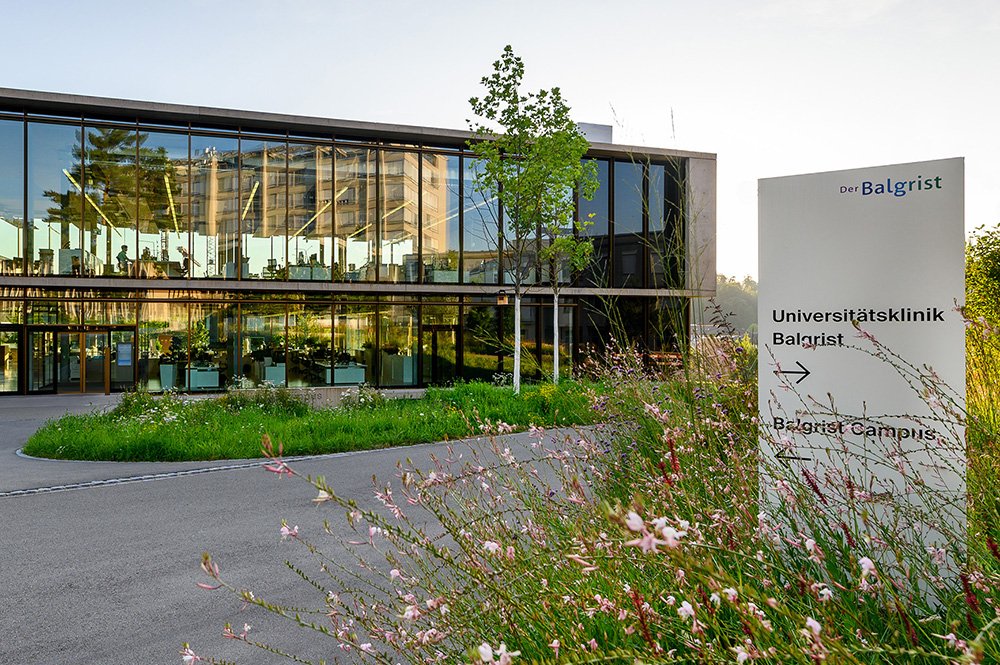 Moderner Gebäudekomplex mit Glasfassade, umgeben von grünen Pflanzen und einem Weg, mit einem Wegweiser, der auf die Universitätsklinik Balgrist und den Balgrist Campus hinweist.