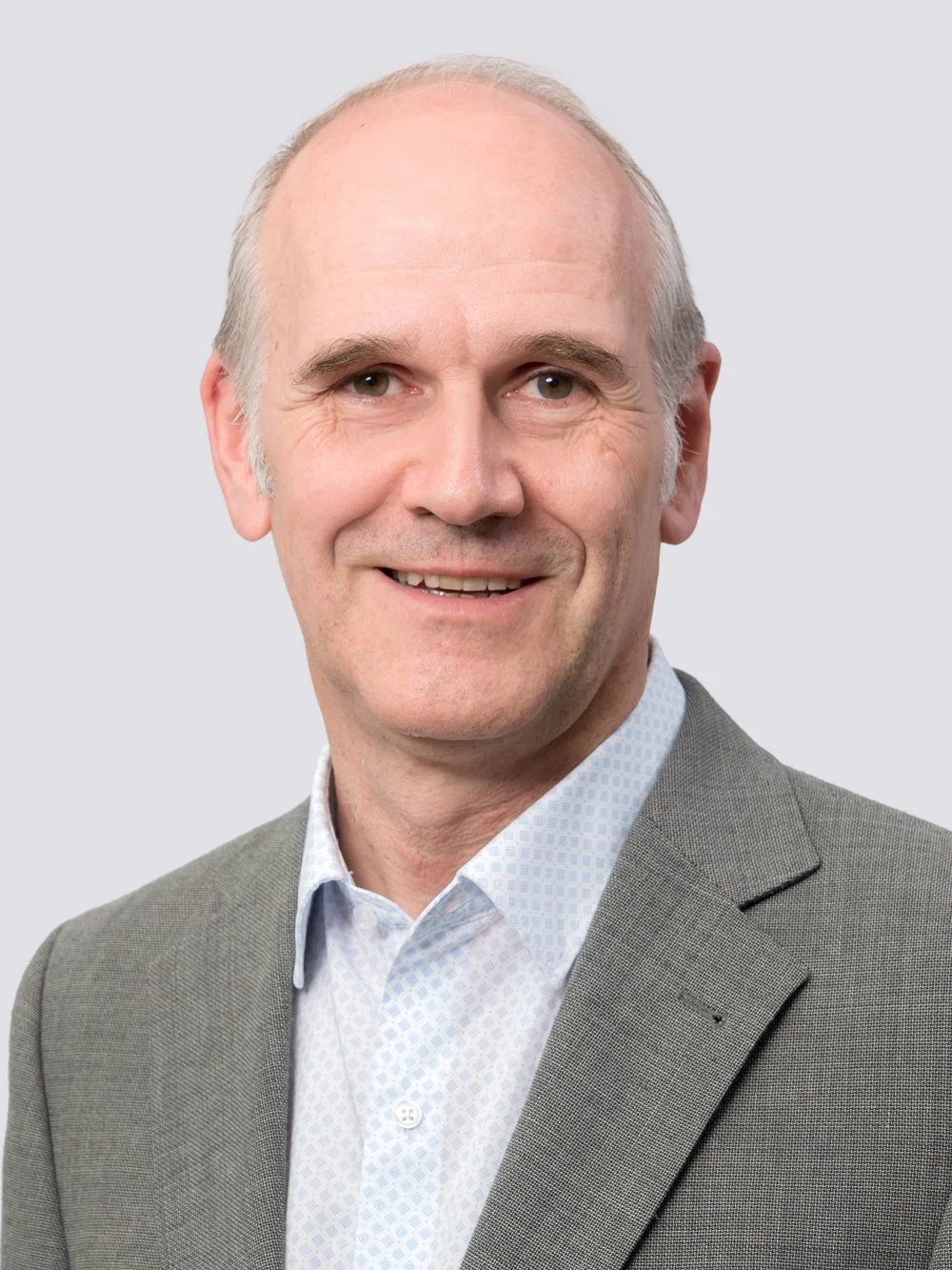 Porträt eines lächelnden Mannes in einem grauen Anzug und weißem Hemd gegen hellgrauen Hintergrund.