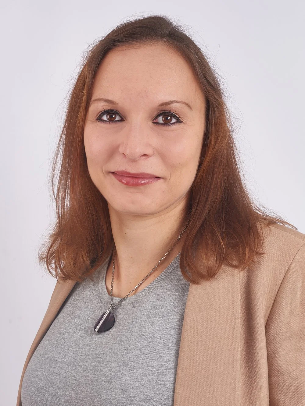 Porträt einer Frau mit braunen Haaren, grauem T-Shirt und beige Blazer vor weißem Hintergrund.