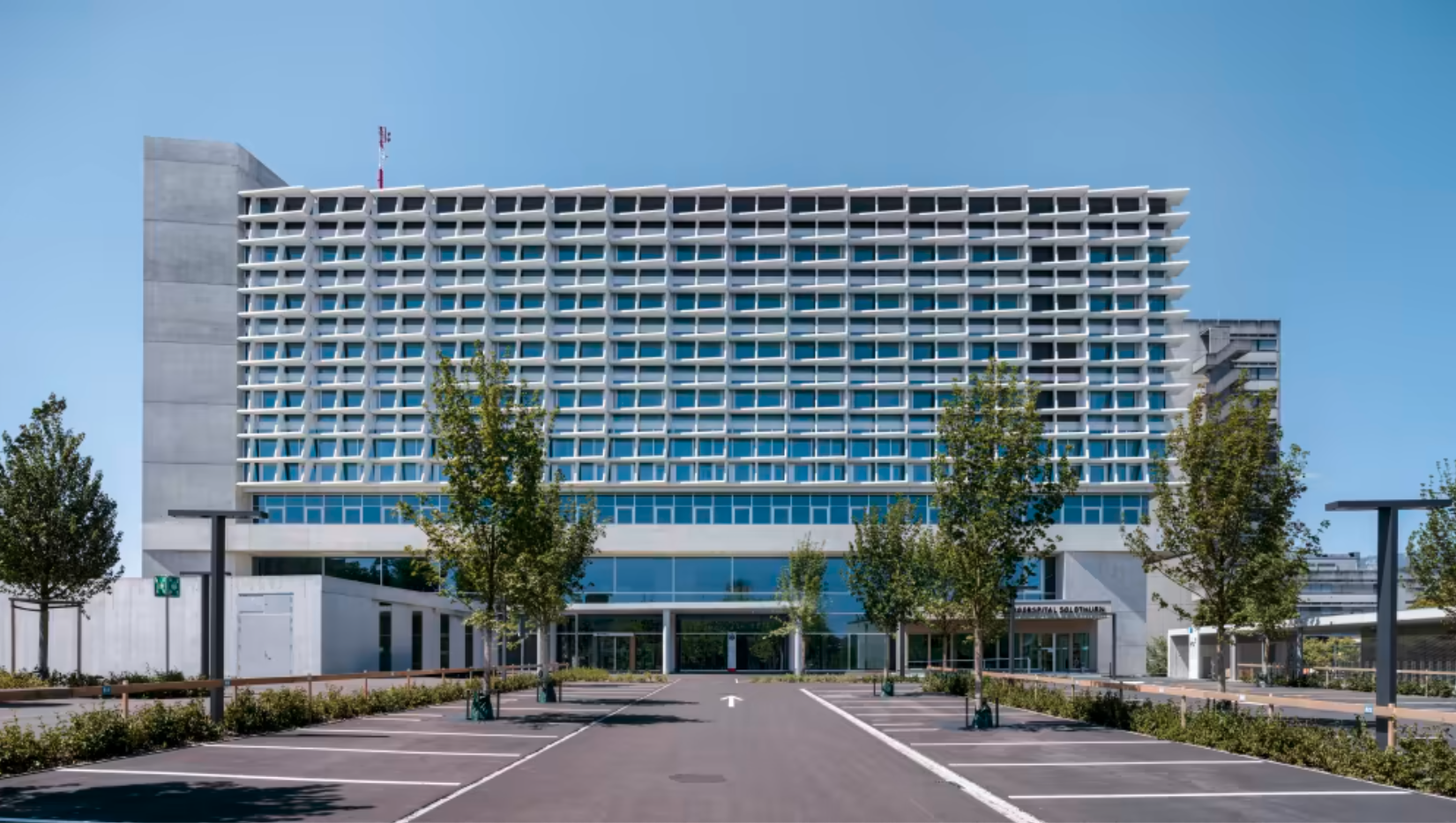 Modernes Krankenhausgebäude mit Parkplätzen und Bäumen vor dem Eingang.