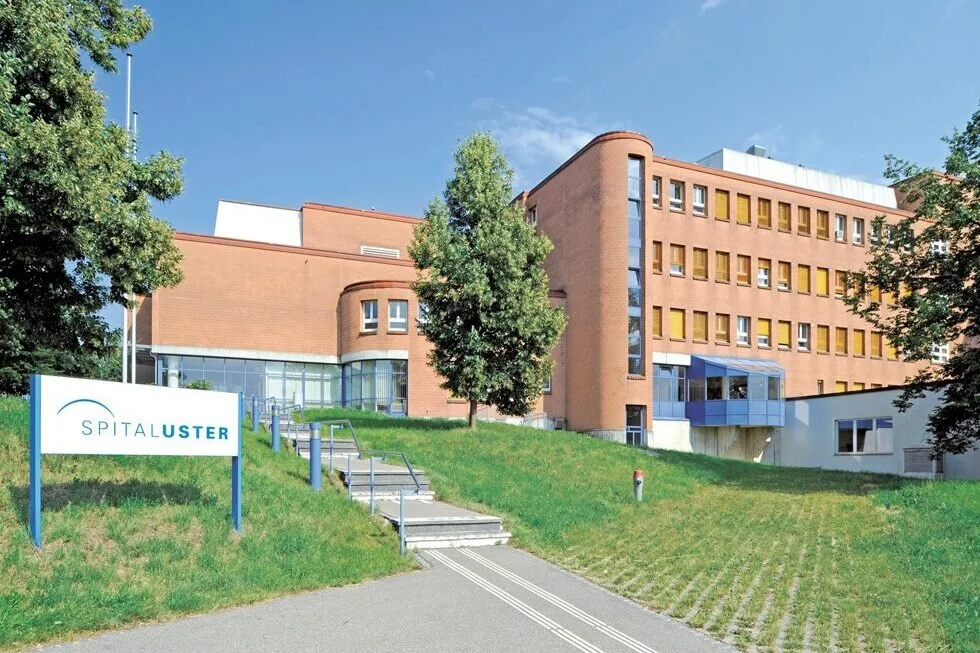 Außenansicht eines Krankenhauses namens "Spital Luster" mit einem gepflegten grünen Rasen, Bäumen und einem blauen Himmel.