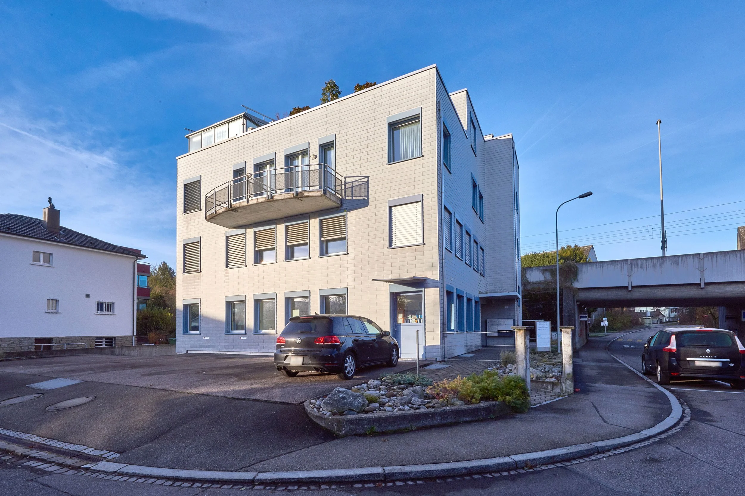 Ein modernes dreistöckiges Gebäude mit weißen Ziegelwänden, mehreren fenster, einem Balkon an der oberen Etage und einem geparkten schwarzen Auto davor. Im Vordergrund eine kleine Grünfläche mit Steinen und Pflanzen, im Hintergrund eine Straße und ein überquerendes Brücke bei Tageslicht.