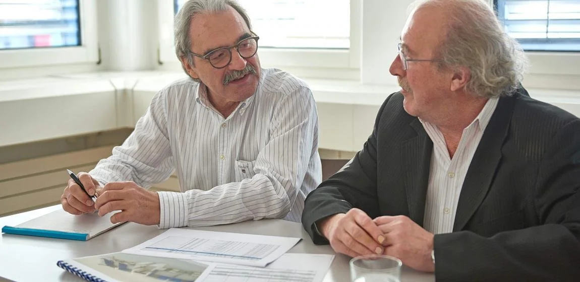Zwei alte Männer sitzen in einem Büro, führen ein Gespräch und lachen miteinander.