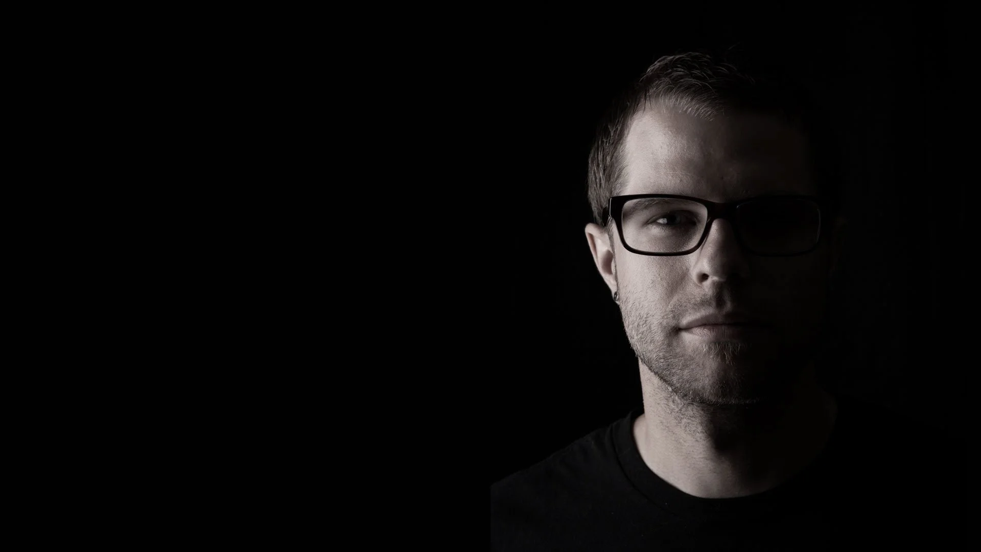 A young man with light skin, short brown hair, wearing black glasses and a black shirt, posed against a dark background with minimal lighting on one side of his face.