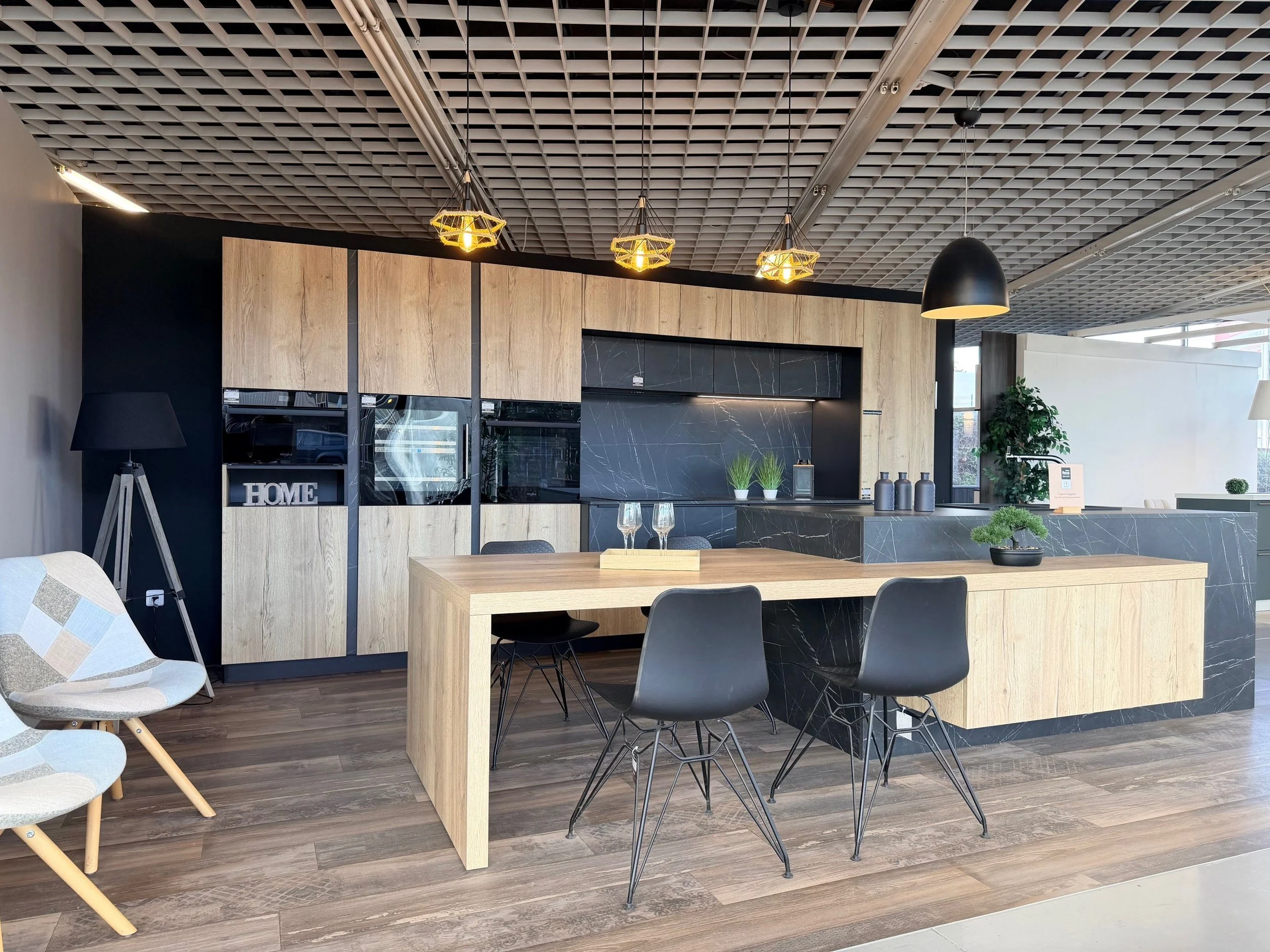 Une cuisine moderne avec un plan de travail en marbre noir, une table en bois clair, des chaises en plastique noir, et des luminaires suspendus au plafond en grille.