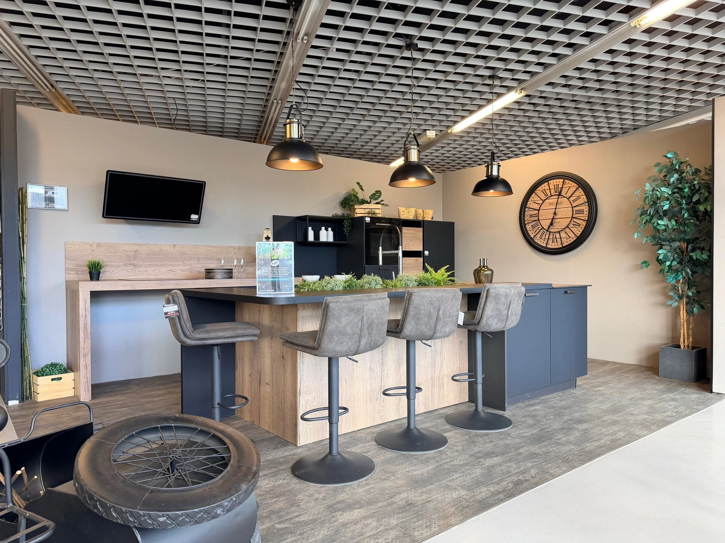 Cuisine moderne avec un plan de travail en bois, quatre chaises en velours gris, un grand mur avec une horloge ancienne en bois, un mur en plâtre blanc, un téléviseur à écran plat au mur, et une grande plante en pot à droite.