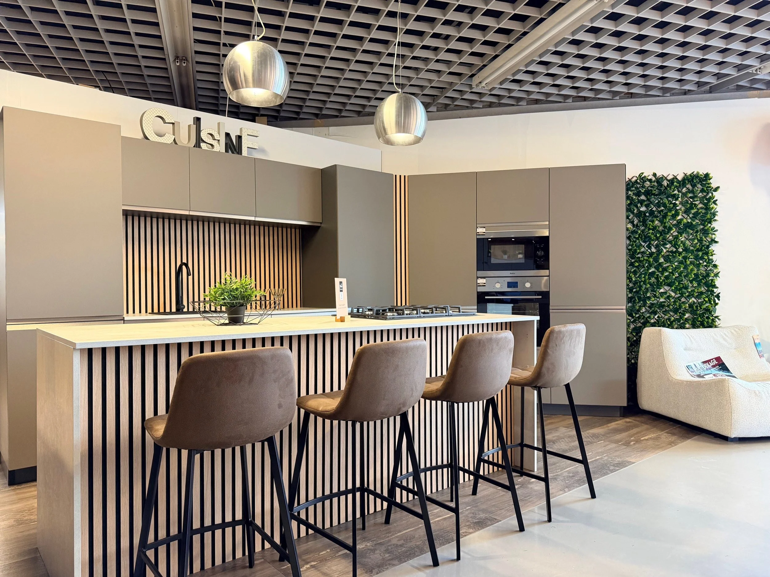 Cuisine moderne avec îlot central en bois et tabourets en velours beige, murs gris, lumière suspendue en métal, décoration végétale, coin salon avec canapé blanc.