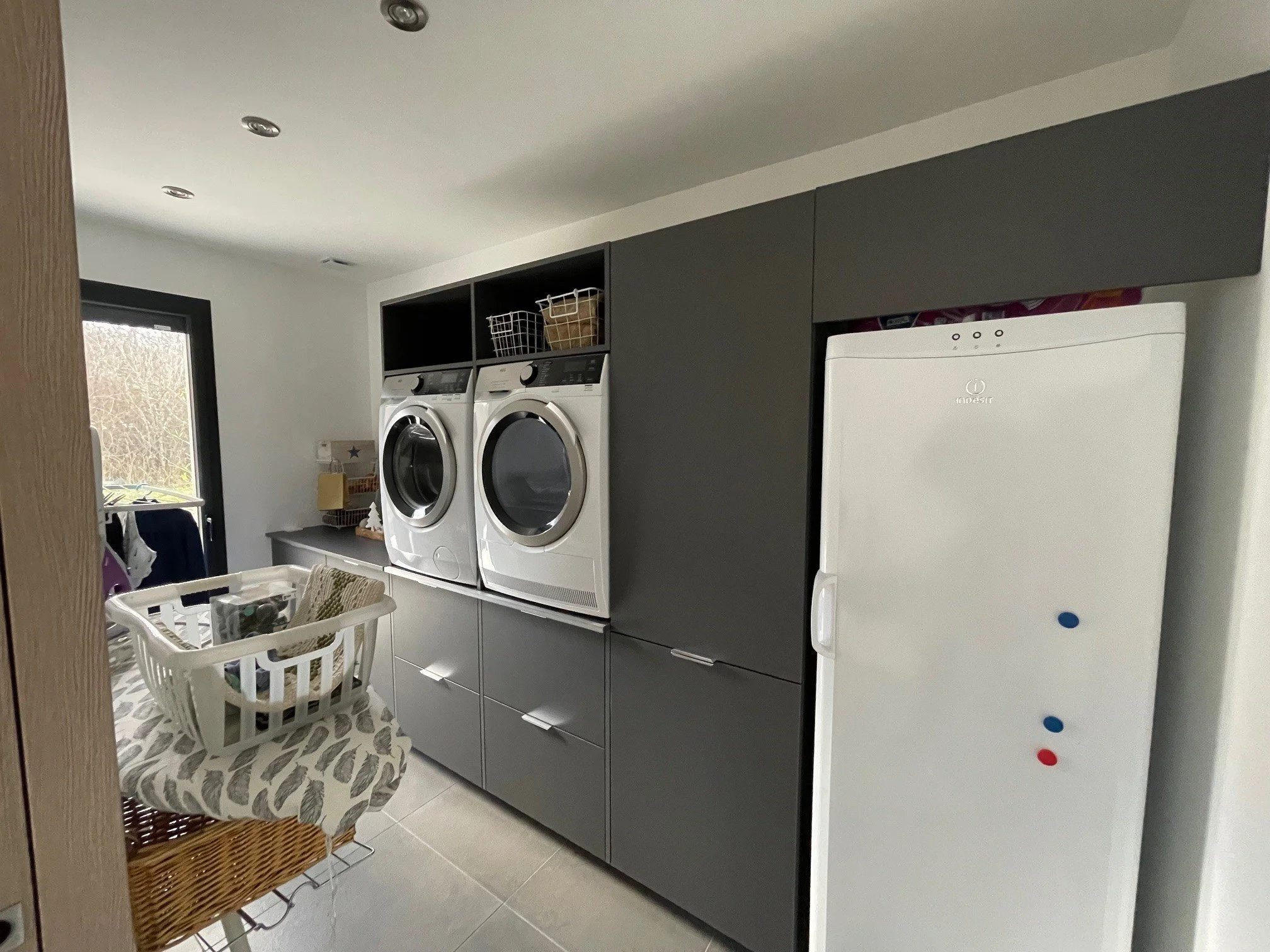 Buanderie avec lave-linge, sèche-linge, réfrigérateur blanc, armoire grise, panier à linge, porte-fenêtre et étagères.