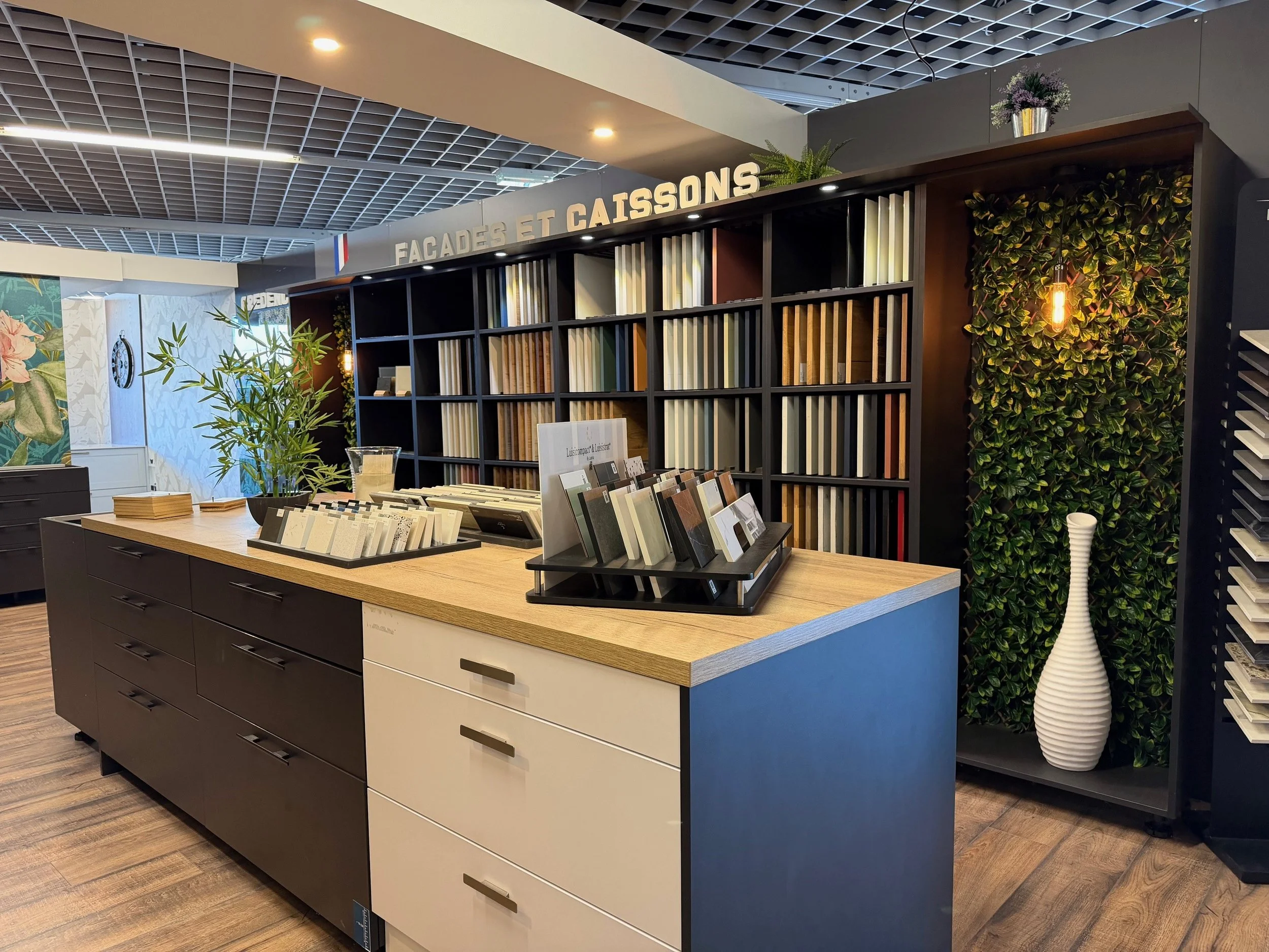 Espace d'exposition avec un comptoir en bois et des étagères remplies de matériaux ou échantillons, décoré avec des plantes vertes et une affiche indiquant 'Façades et Caissons'.
